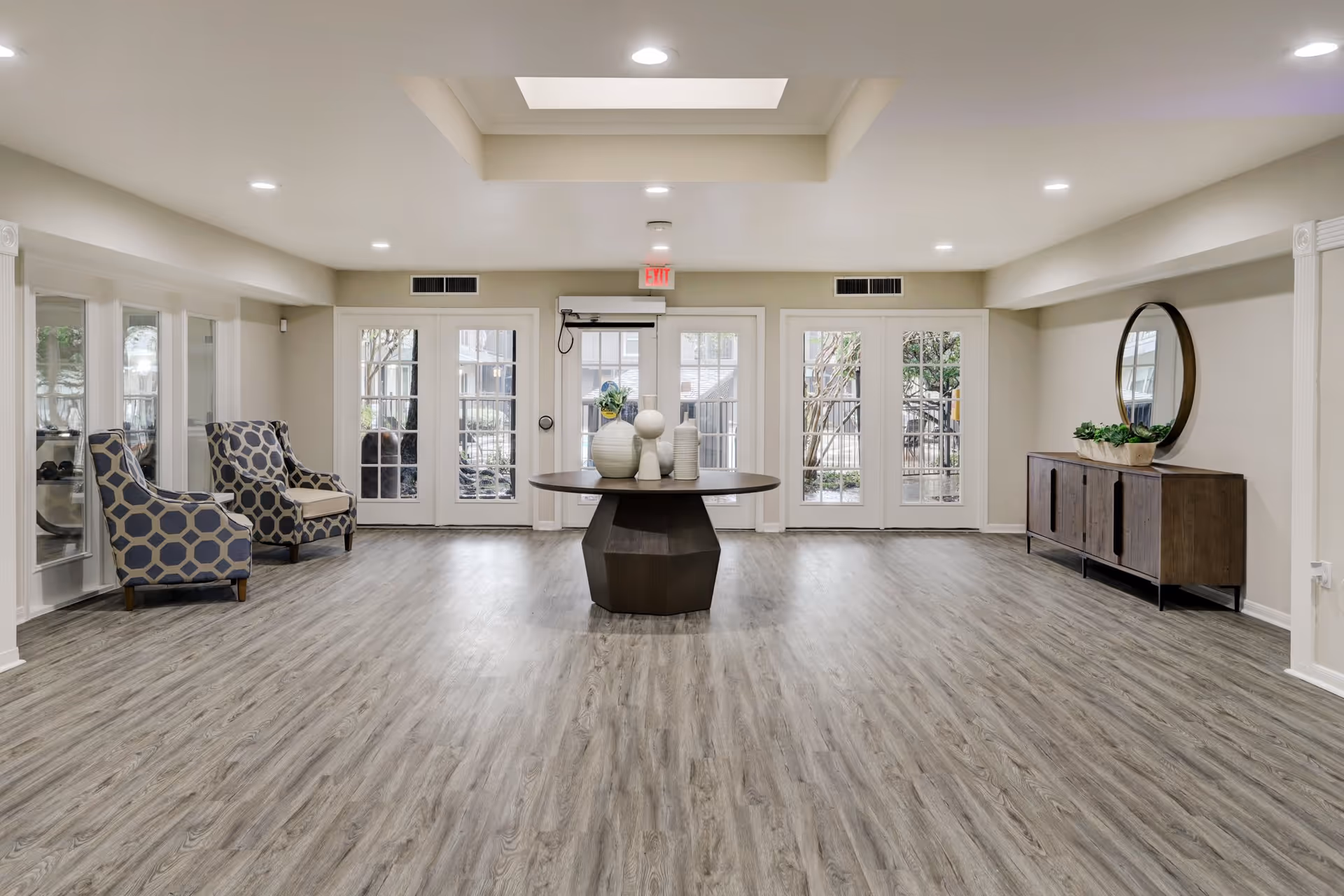 A spacious, well-lit interior room with light wood flooring and beige walls. The room features a round wooden table in the center with decorative vases, two patterned armchairs on the left side, and a wooden sideboard with a round mirror and potted plants on the right side. Large glass doors at the back provide a view of an outdoor area with trees and a fence.