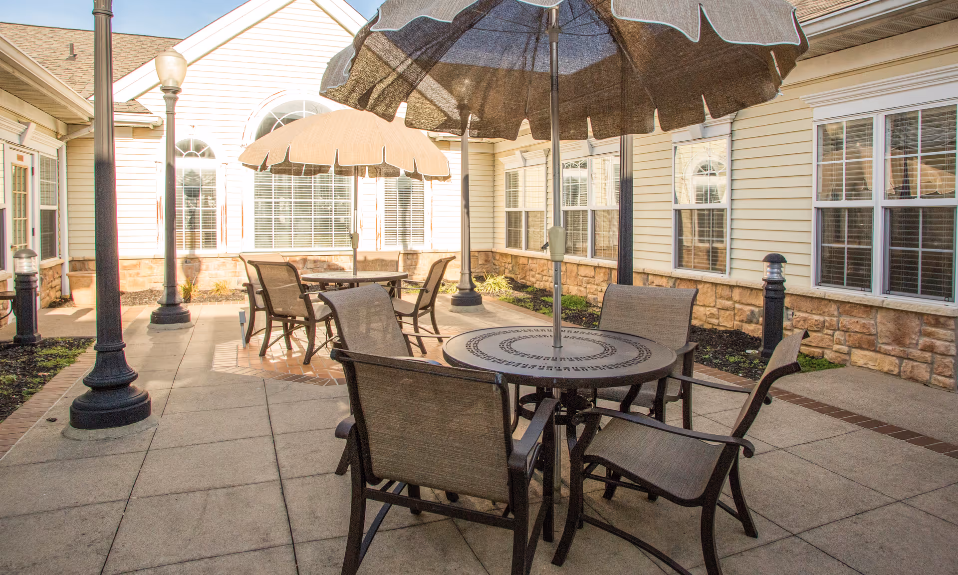 Outdoor patio area with round tables and chairs under large umbrellas, surrounded by a building with multiple windows and stone siding at the base.