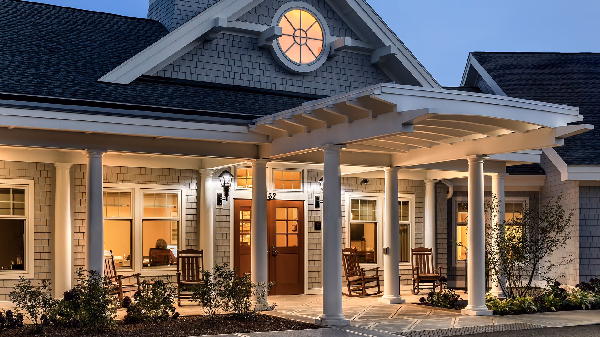 Exterior view of a senior living facility entrance at dusk with a covered porch supported by white columns. The building has gray shingles, warm interior lighting visible through the windows, and several wooden rocking chairs on the porch. There are small bushes and plants around the entrance area.