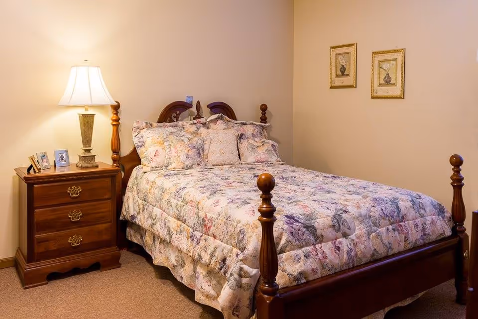A cozy bedroom with a wooden bed frame and a floral-patterned bedspread and pillows. Next to the bed is a wooden nightstand with three drawers, a table lamp, and framed photos. Two framed floral pictures hang on the beige wall above the bed.