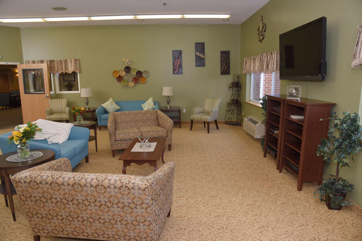 A cozy living room with green walls and beige patterned carpet. The room features multiple seating options including a patterned sofa, a blue loveseat, and striped armchairs. There are side tables with lamps and flower vases, a wooden coffee table with a decorative piece, a flat-screen TV mounted on the wall above wooden shelves, and wall art decorations. A window with valances lets in natural light.