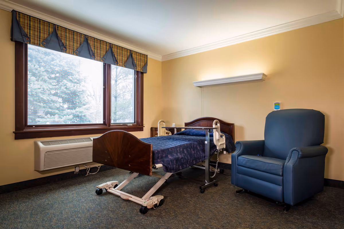 A simple assisted living bedroom with a hospital-style bed covered in a dark blue patterned blanket, a large blue recliner chair, a window with blinds and a plaid valance, beige walls, and a carpeted floor.