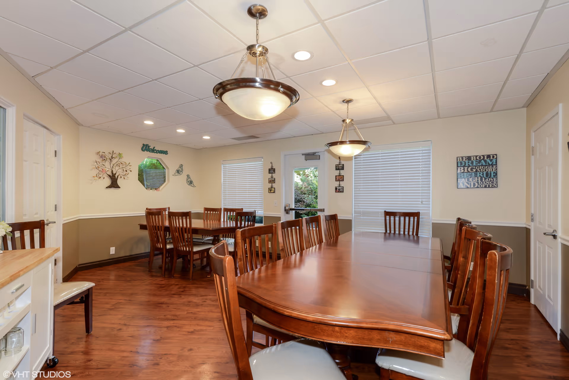 A dining room with two large wooden tables surrounded by wooden chairs with cushioned seats. The room has wood flooring, beige and brown walls, and a white ceiling with recessed lighting and two hanging light fixtures. There are two windows with blinds, a door leading outside, and decorative wall art including a tree, birds, a welcome sign, and an inspirational quote.