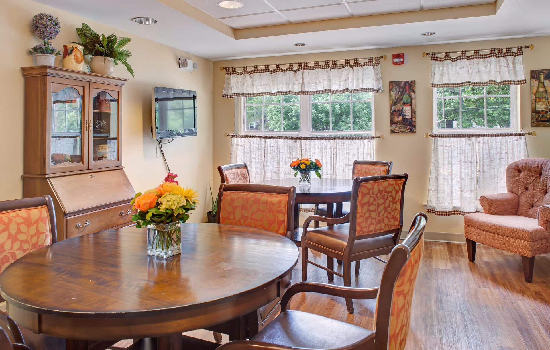A cozy dining room with two round wooden tables, each adorned with a vase of colorful flowers. The room features several cushioned chairs with patterned upholstery, a wooden cabinet with plants on top, a wall-mounted TV, and large windows with white curtains letting in natural light. There is also a comfortable upholstered armchair near the windows.