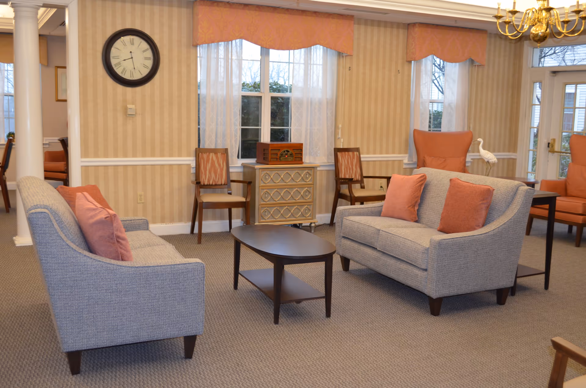 A cozy living room area with two gray sofas adorned with orange throw pillows facing each other across a dark wooden coffee table. The room features beige striped wallpaper, a large wall clock, two wooden chairs, a small decorative cabinet, and large windows with sheer white curtains and orange valances. There is also a chandelier and a decorative white bird statue near the window.