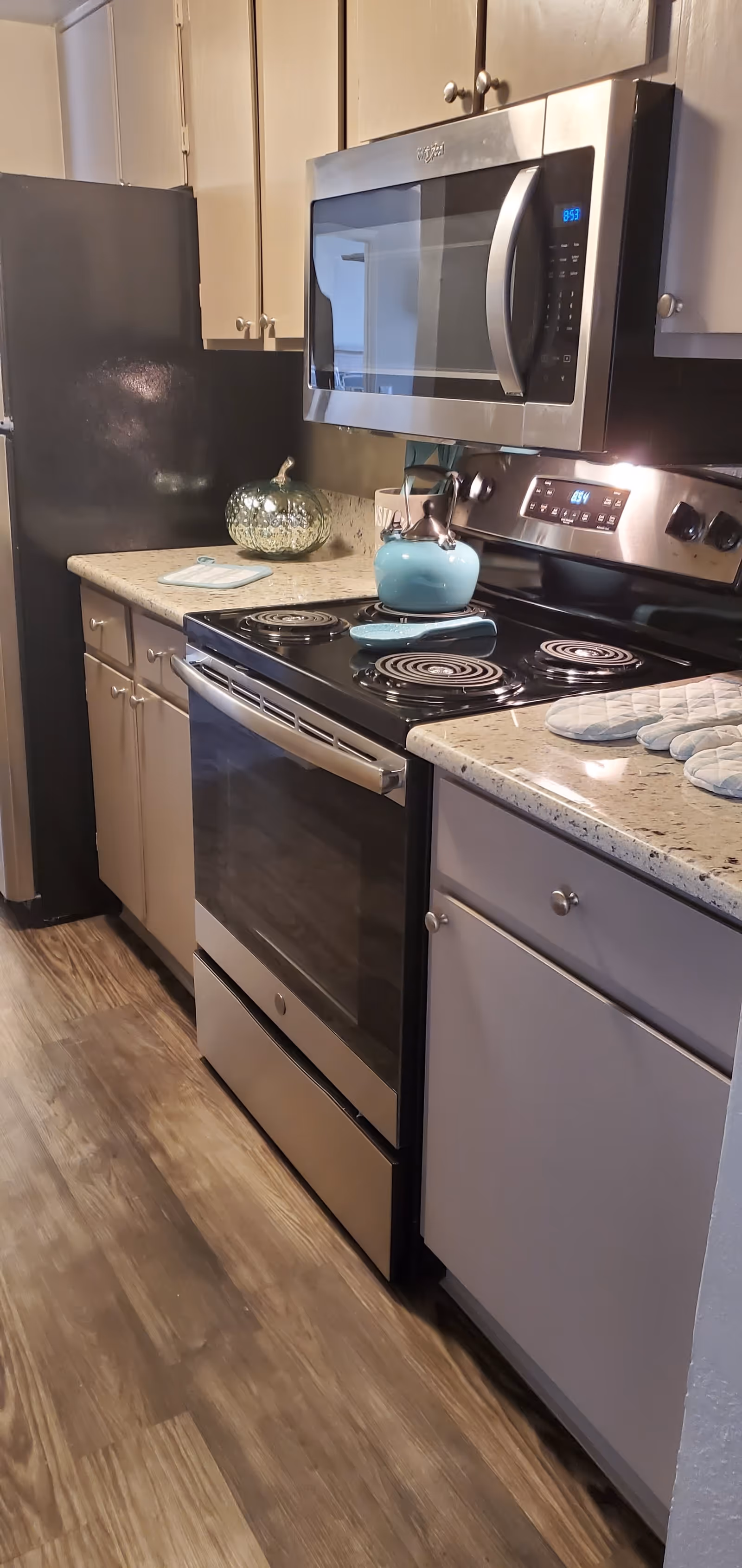 A modern kitchen with a stainless steel electric stove and oven, a matching microwave above it, beige cabinets, a black refrigerator, and a countertop with a decorative glass pumpkin, a blue kettle on the stove, and blue oven mitts.
