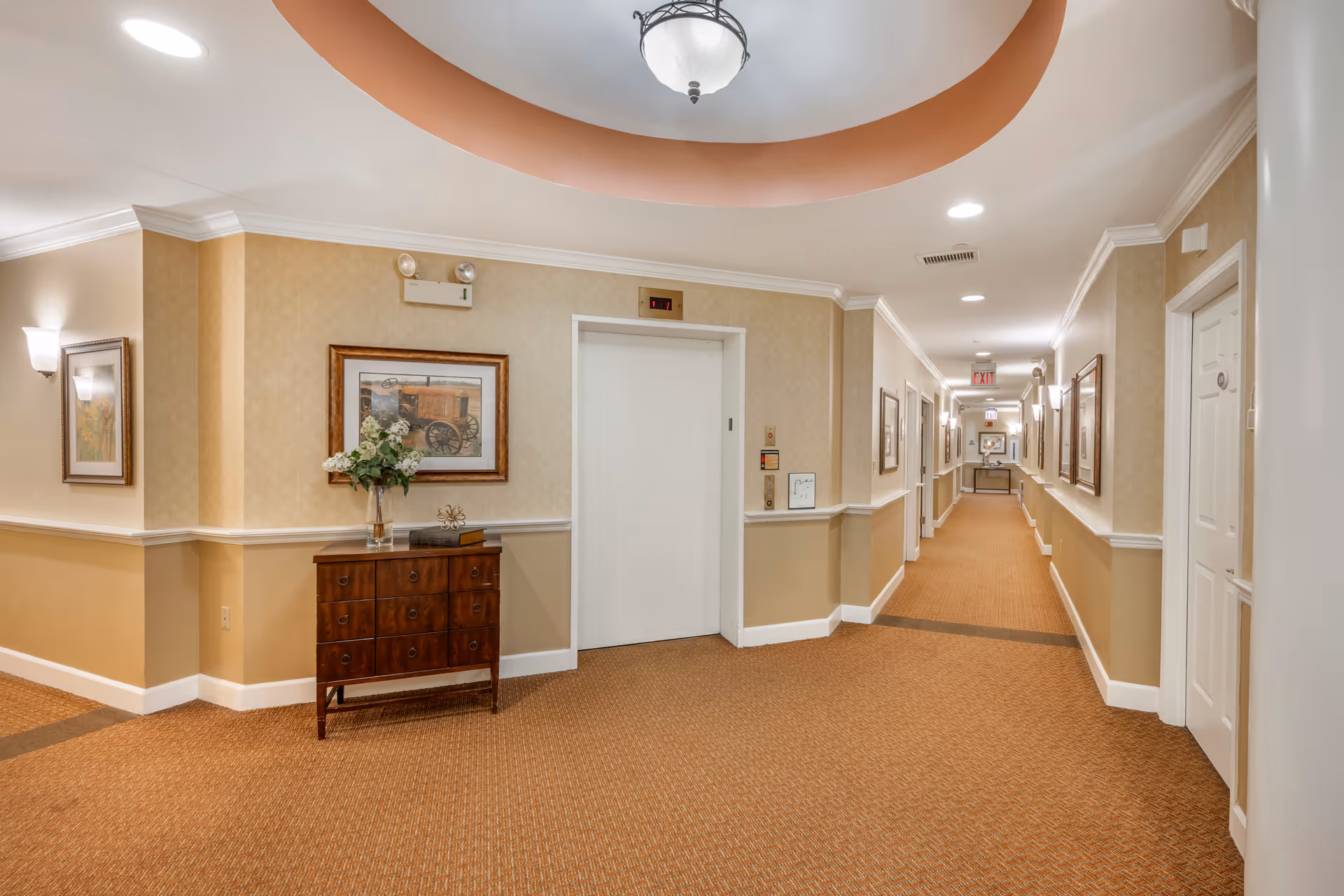 Interior hallway of a senior living facility with beige walls and carpeted floor. There is a white elevator door in the center with a small wooden cabinet to the left holding a vase with flowers and decorative items. The hallway extends to the right with several closed white doors and framed artwork on the walls. Ceiling lights and wall sconces illuminate the space.