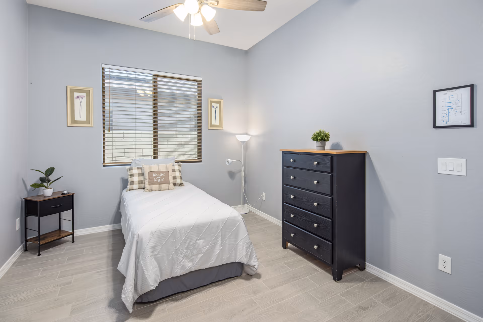 A small, neatly arranged bedroom with a single bed covered in white bedding and a decorative pillow that reads 'thankful & blessed'. The room has light gray walls, a window with wooden blinds, two framed floral pictures on either side of the window, a black nightstand with a small plant and an alarm clock, a black chest of drawers with a small potted plant on top, a floor lamp in the corner, and a ceiling fan with lights.