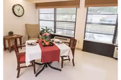 Dining area with a table set with a white cloth and red runner, surrounded by chairs and an armchair beneath large windows with blinds and a wall clock.