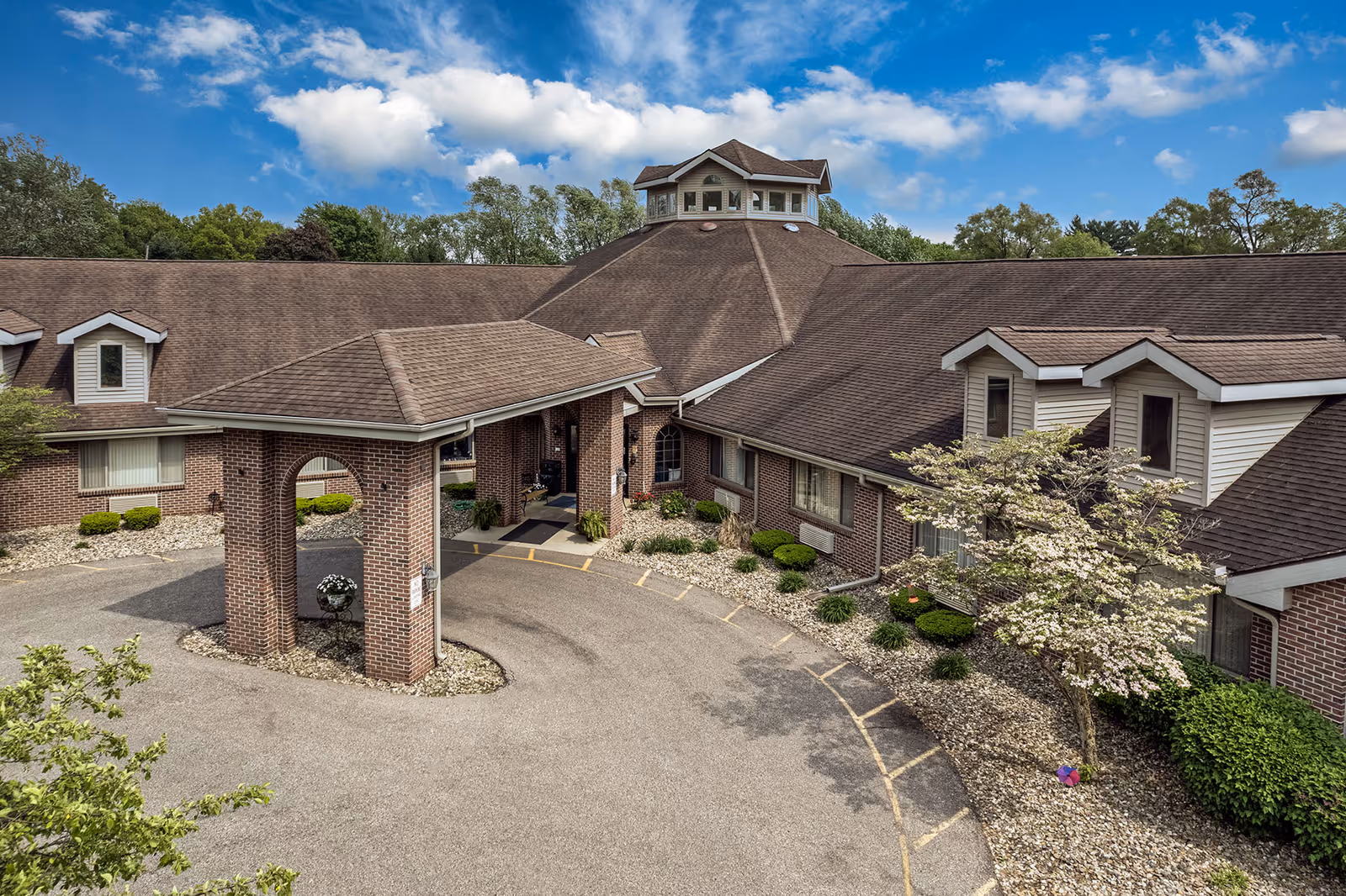 Exterior view of Brentwood at Elkhart Assisted Living facility showing a brick building with a covered entrance driveway, surrounded by landscaping with bushes and a tree, under a partly cloudy blue sky.