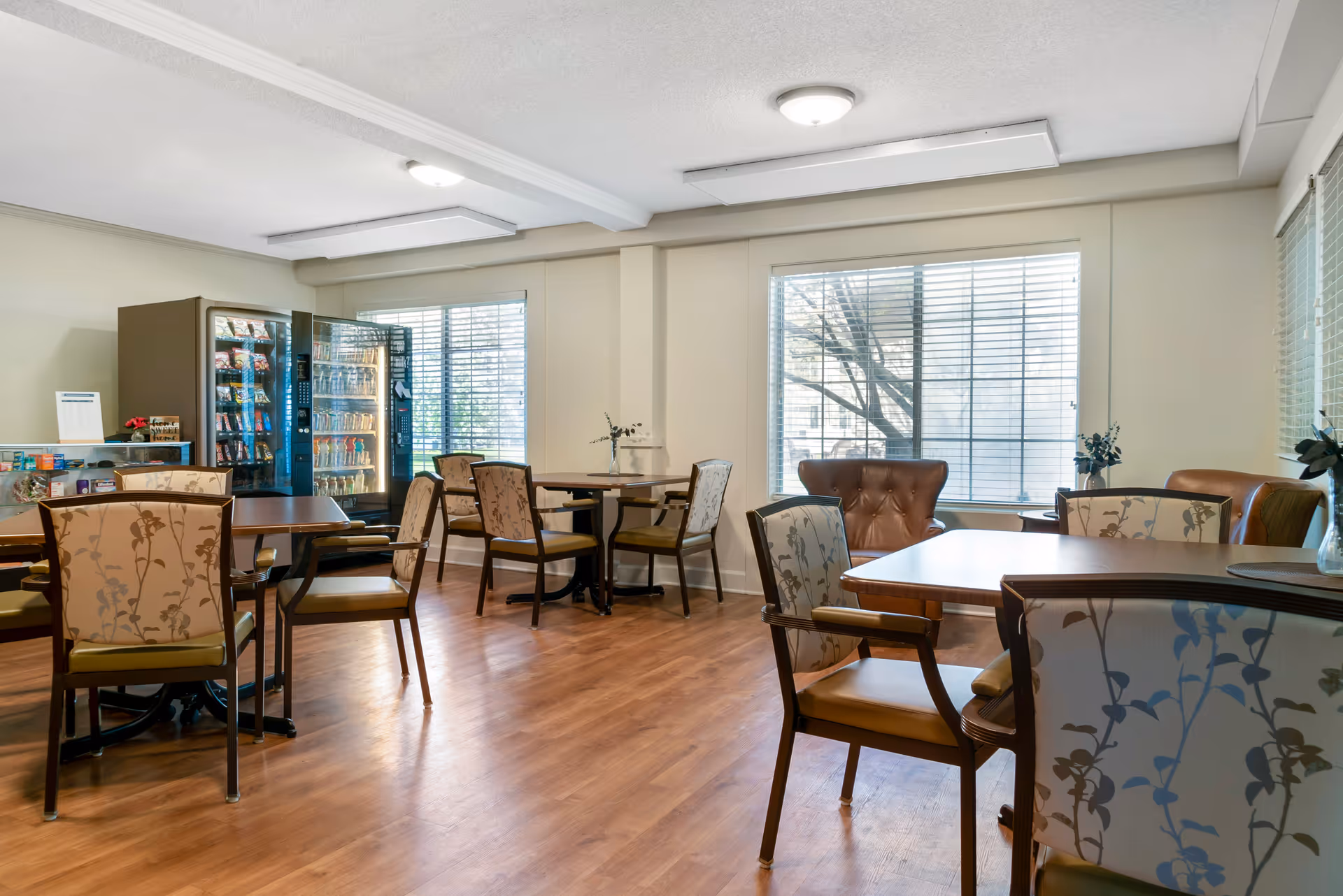 A bright room with wooden flooring featuring several tables and chairs with floral upholstery. Large windows with blinds allow natural light to fill the space. In the corner, there are vending machines stocked with snacks and drinks. The room has a clean, welcoming atmosphere.