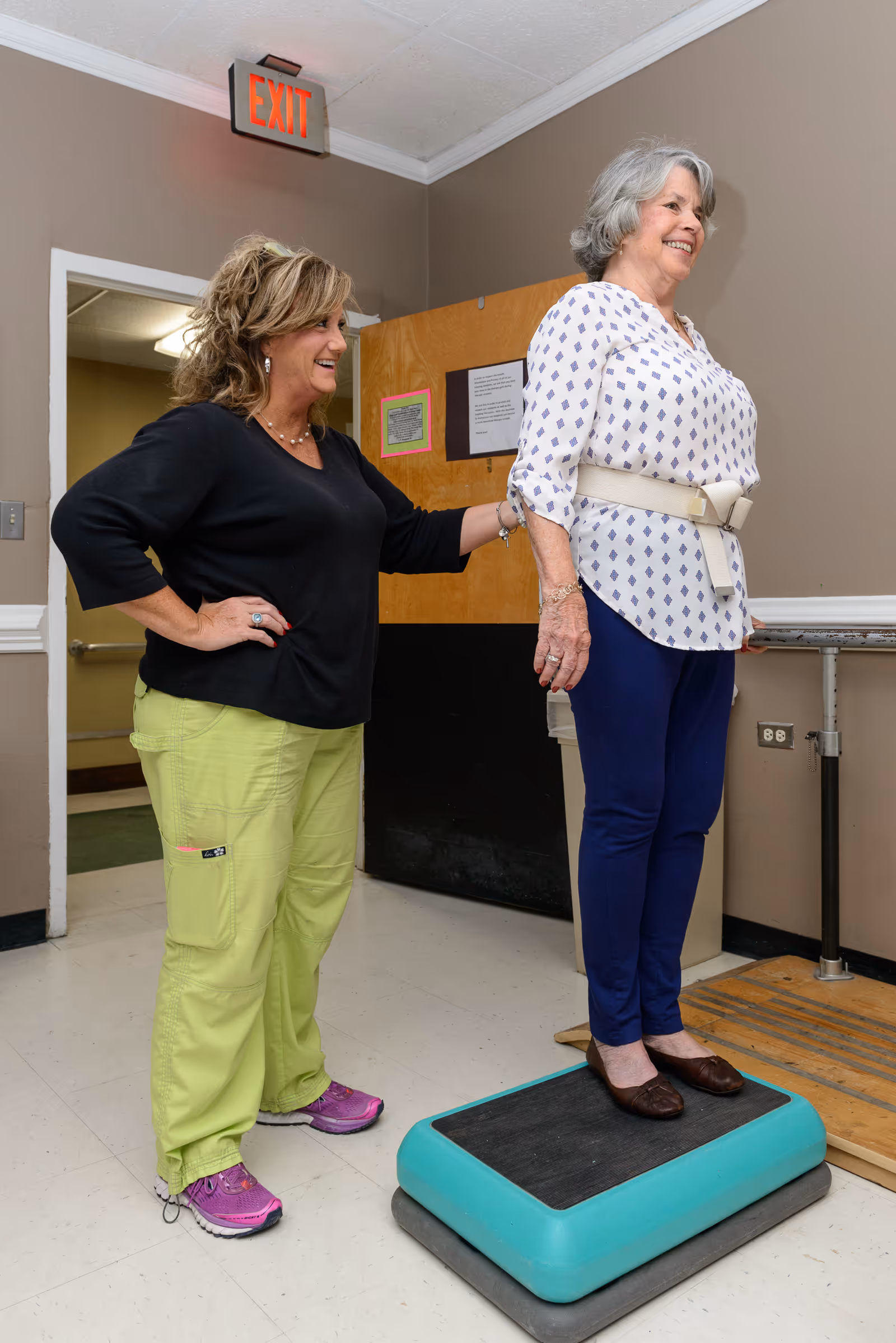 A senior woman stands on a step platform wearing a posture support belt while a caregiver or therapist stands beside her, providing assistance and encouragement in a rehabilitation or therapy room.