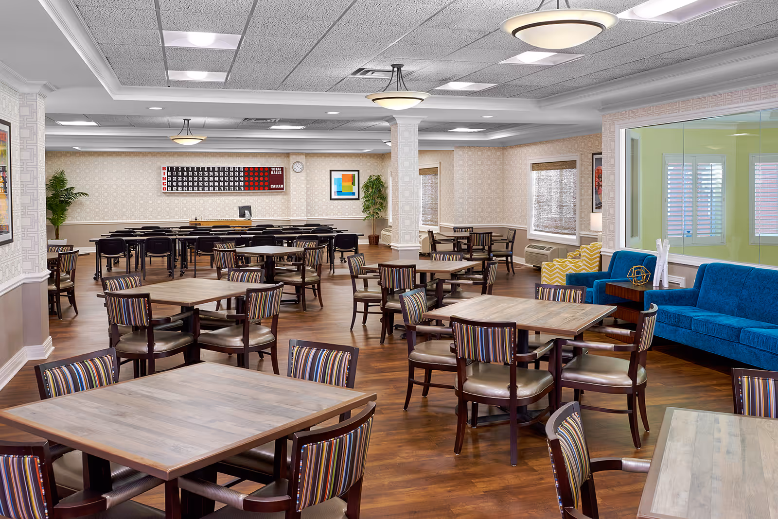 A spacious, well-lit common area with multiple wooden tables and chairs arranged for group seating. The room features a blue couch and a yellow patterned armchair near a large window with white shutters. The walls are decorated with framed artwork and a large bingo scoreboard. The floor is wooden, and the ceiling has recessed lighting and decorative light fixtures.