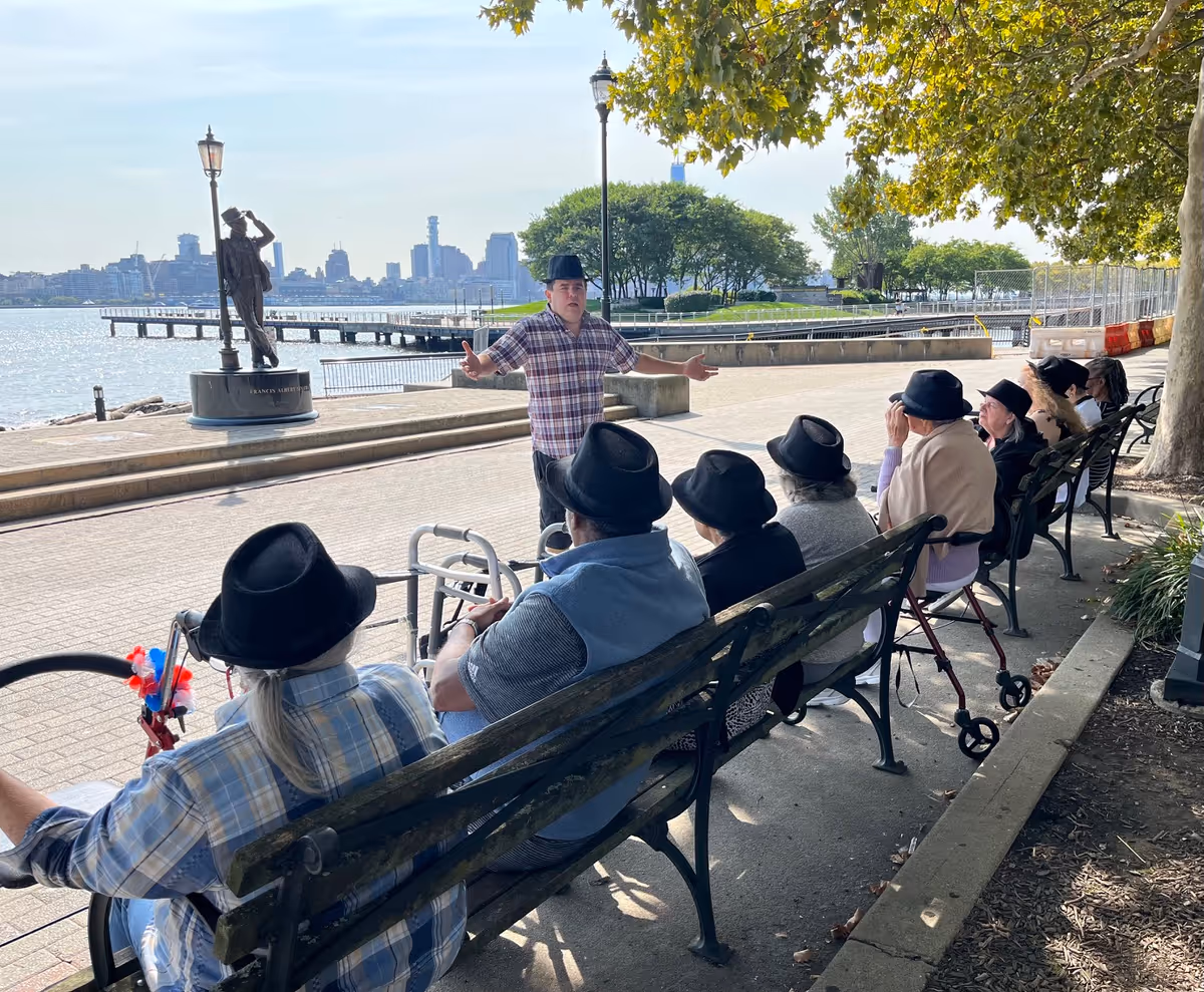 A group of elderly people wearing black hats sitting on benches outdoors near a waterfront, listening to a man in a plaid shirt and black hat who is standing and gesturing in front of them. There is a statue, lampposts, trees, and a city skyline in the background.