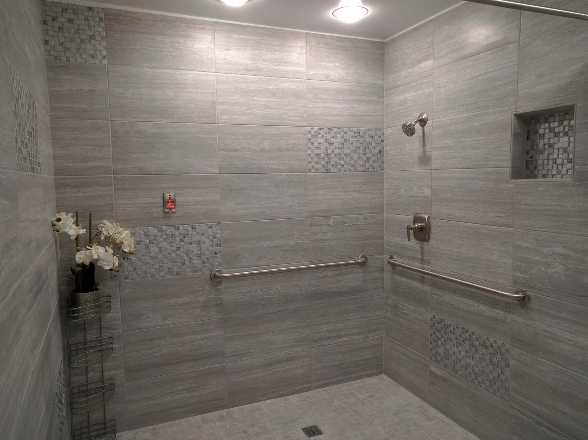 A spacious, tiled shower area with gray tiles and mosaic tile accents. The shower features two stainless steel grab bars on adjacent walls, a wall-mounted showerhead, and a built-in recessed shelf with mosaic tiles. A metal rack with white orchids is placed in the corner. The ceiling has a round light fixture.