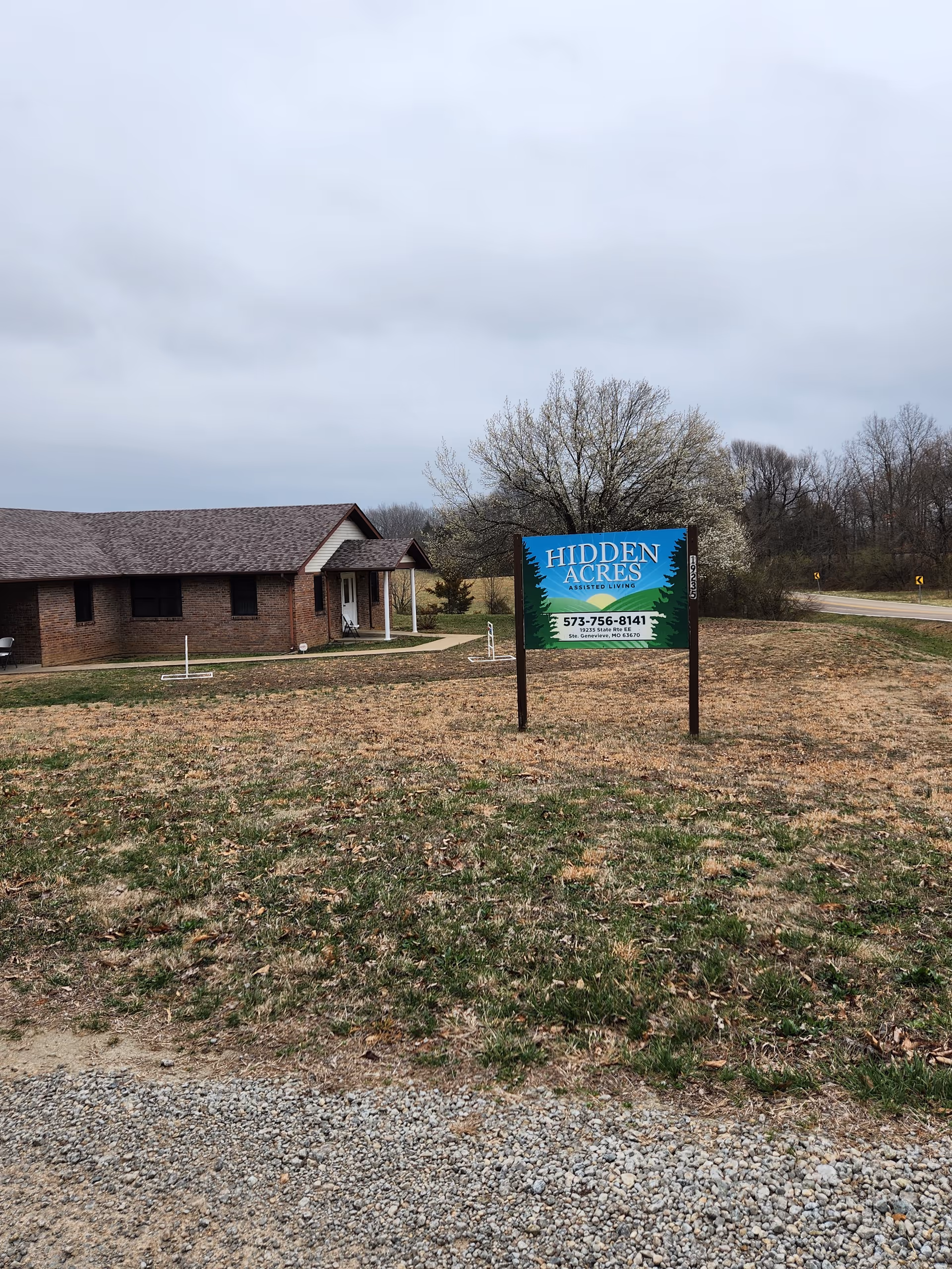 Front exterior of a brick assisted living building with a 'Hidden Acres' sign on the lawn.
