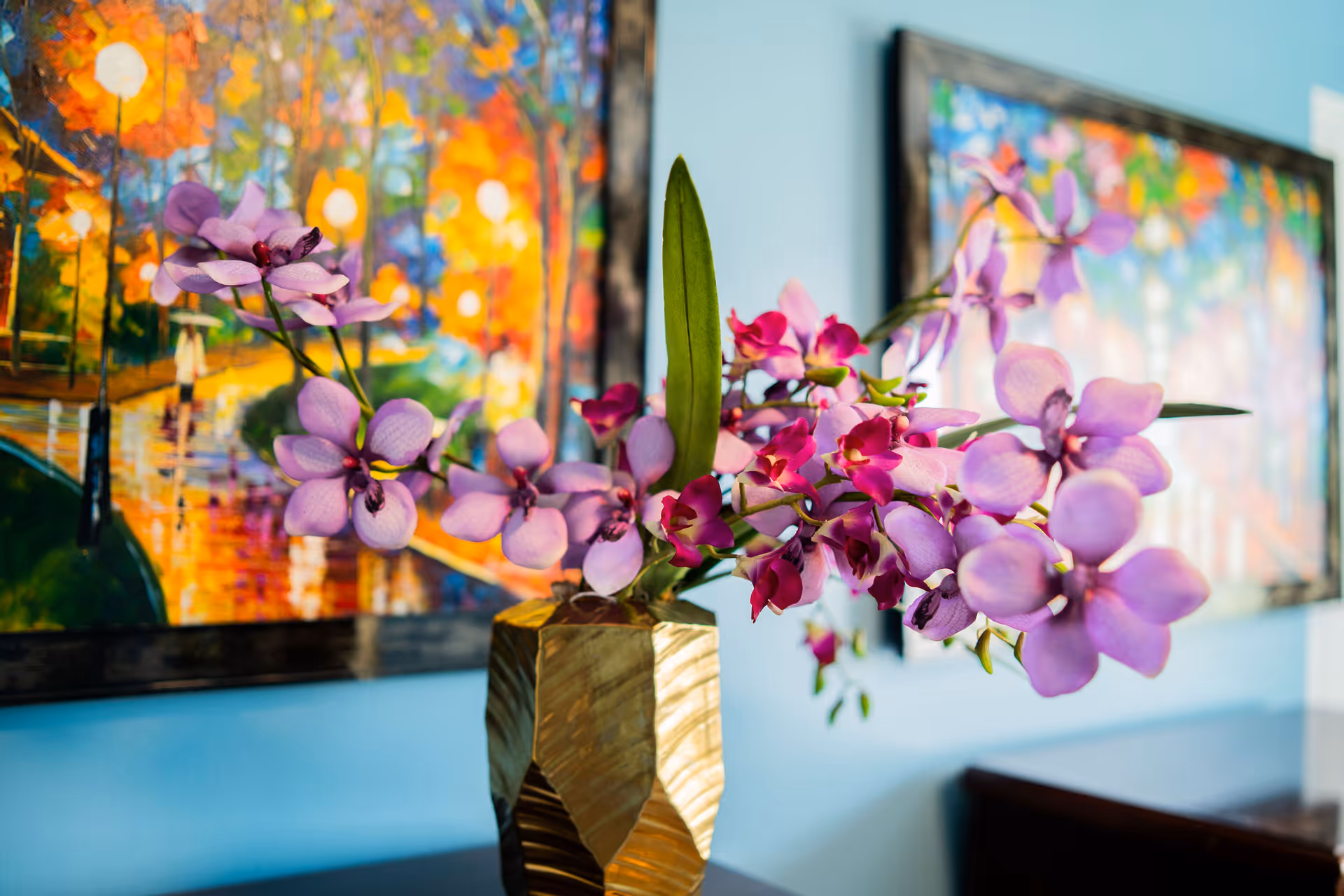 A gold vase filled with purple orchids placed in front of colorful framed paintings on a blue wall.