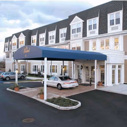 Front entrance of a multi-story assisted living building with a blue awning, driveway islands, and parked cars.