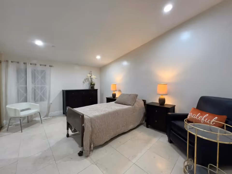 A neatly arranged bedroom with a single bed covered in a beige bedspread, two bedside tables each with a lit lamp, a black dresser with a flower vase on top, a white chair near a window with sheer curtains, and a black armchair with an orange pillow that says 'grateful' next to a small round glass table.