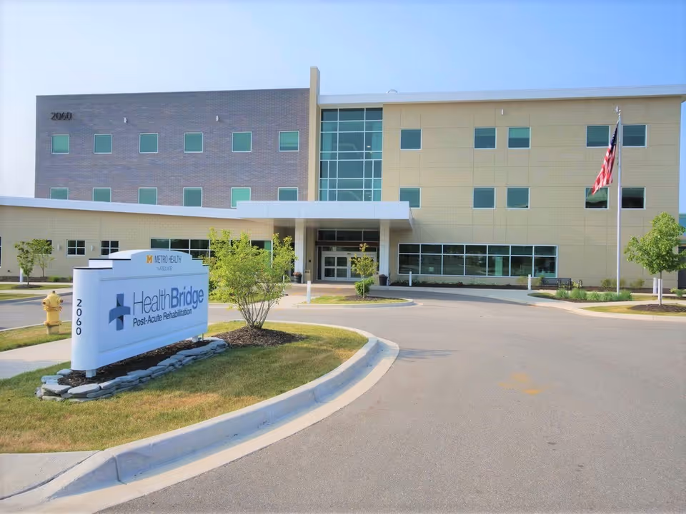Exterior view of a modern three-story post-acute rehabilitation center building with a sign in front that reads 'HealthBridge Post-Acute Rehabilitation' and 'Metro Health'. The building has multiple windows, a covered entrance, a flagpole with an American flag, and landscaped greenery around the driveway.