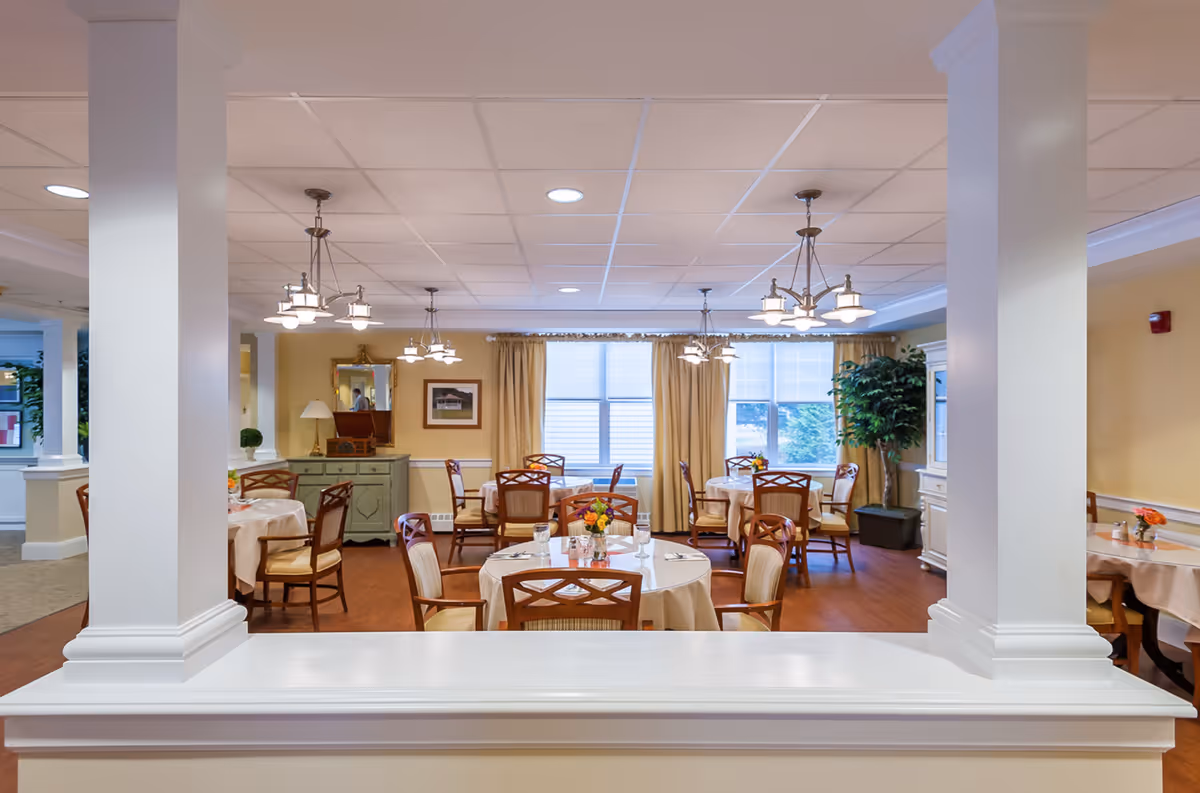 A bright and inviting dining room with round tables covered in beige tablecloths, each set with glasses, napkins, and small flower arrangements. Wooden chairs with cushioned seats surround the tables. The room features large windows with beige curtains, multiple ceiling light fixtures, and decorative plants. The walls are painted a soft yellow, and there are white columns and trim in the foreground.