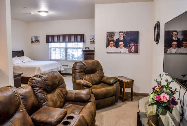 A cozy living area with two brown leather recliners facing a large flat-screen TV mounted on the wall. A small wooden side table with a marble top sits between the recliners. In the background, there is a bedroom with a bed covered in white bedding, a window with a valance, and a dresser. The walls are decorated with framed family photos and a round mirror. A vase with colorful flowers is placed on a glass table near the TV.