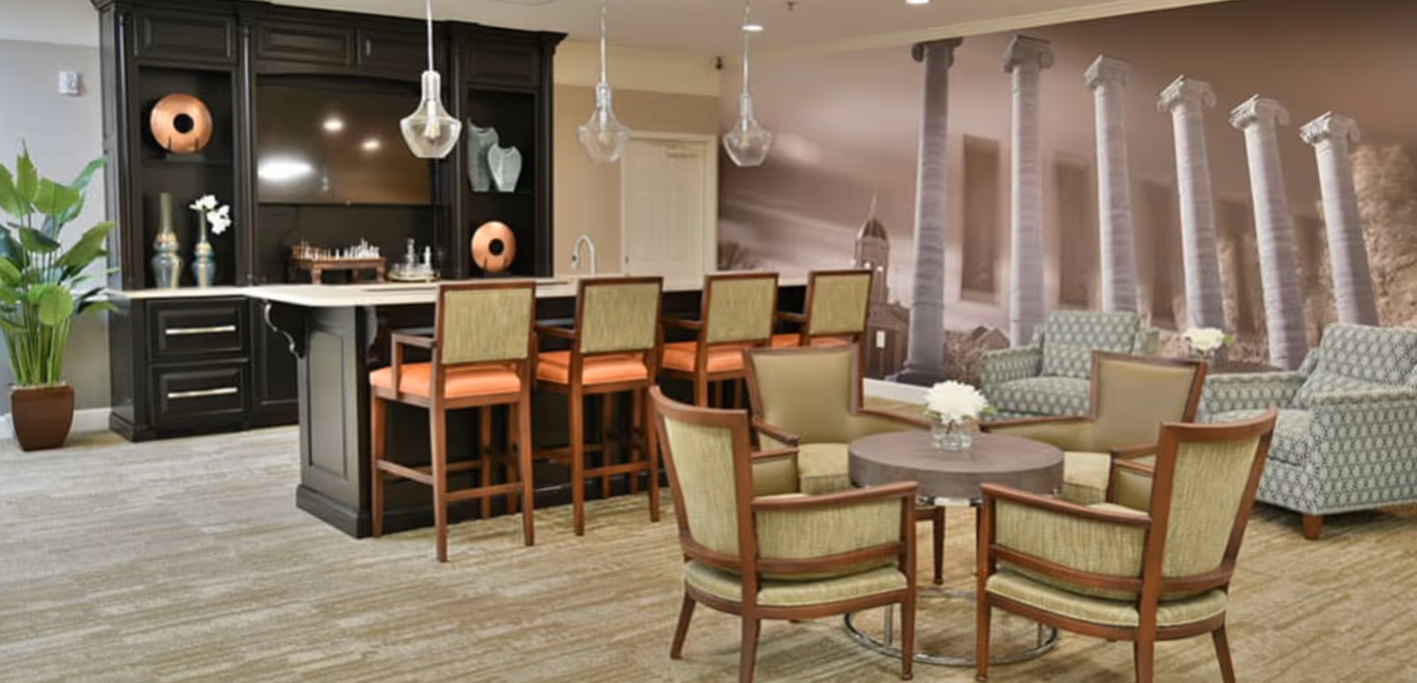 A cozy common area with a seating arrangement including four green cushioned chairs around a round wooden table with a small flower vase. Behind the seating area, there are two patterned armchairs. To the left, there is a kitchen island with four high chairs with orange cushions. The back wall has dark cabinetry with decorative items and a large TV. The wall features a mural of classical columns and a building. A large potted plant is placed on the left side of the room.