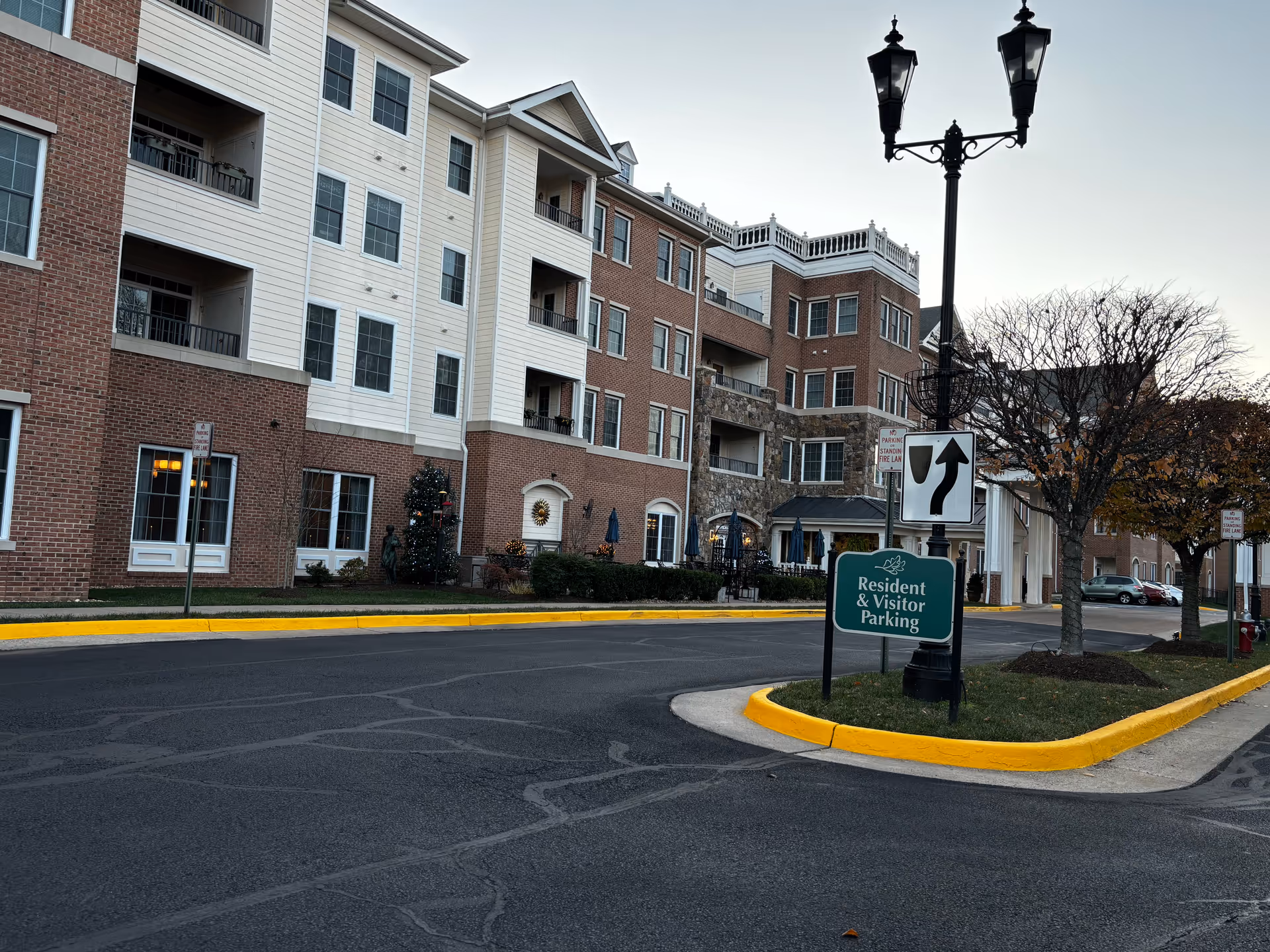 Exterior view of a multi-story senior living facility building with brick and siding facade, balconies, and large windows. In front, there is a street with a green sign indicating Resident & Visitor Parking, a street lamp, and some trees with autumn leaves.