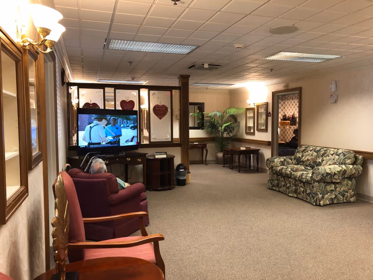 A common-area lounge with upholstered chairs and a floral sofa, a TV playing a program, and an elderly resident seated watching.