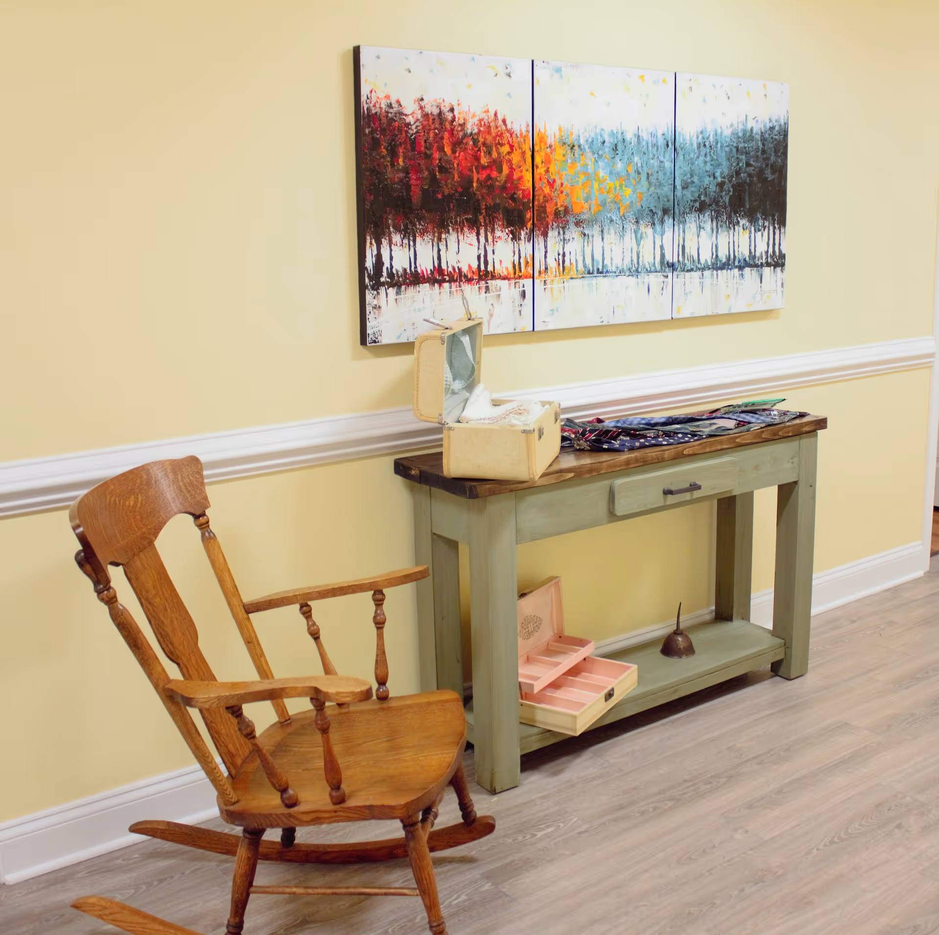 A cozy interior corner featuring a wooden rocking chair next to a green console table with a drawer. On the table are various items including a small open box and a collection of ties. Above the table hangs a three-panel abstract painting depicting a row of trees in autumn colors transitioning to cooler tones.
