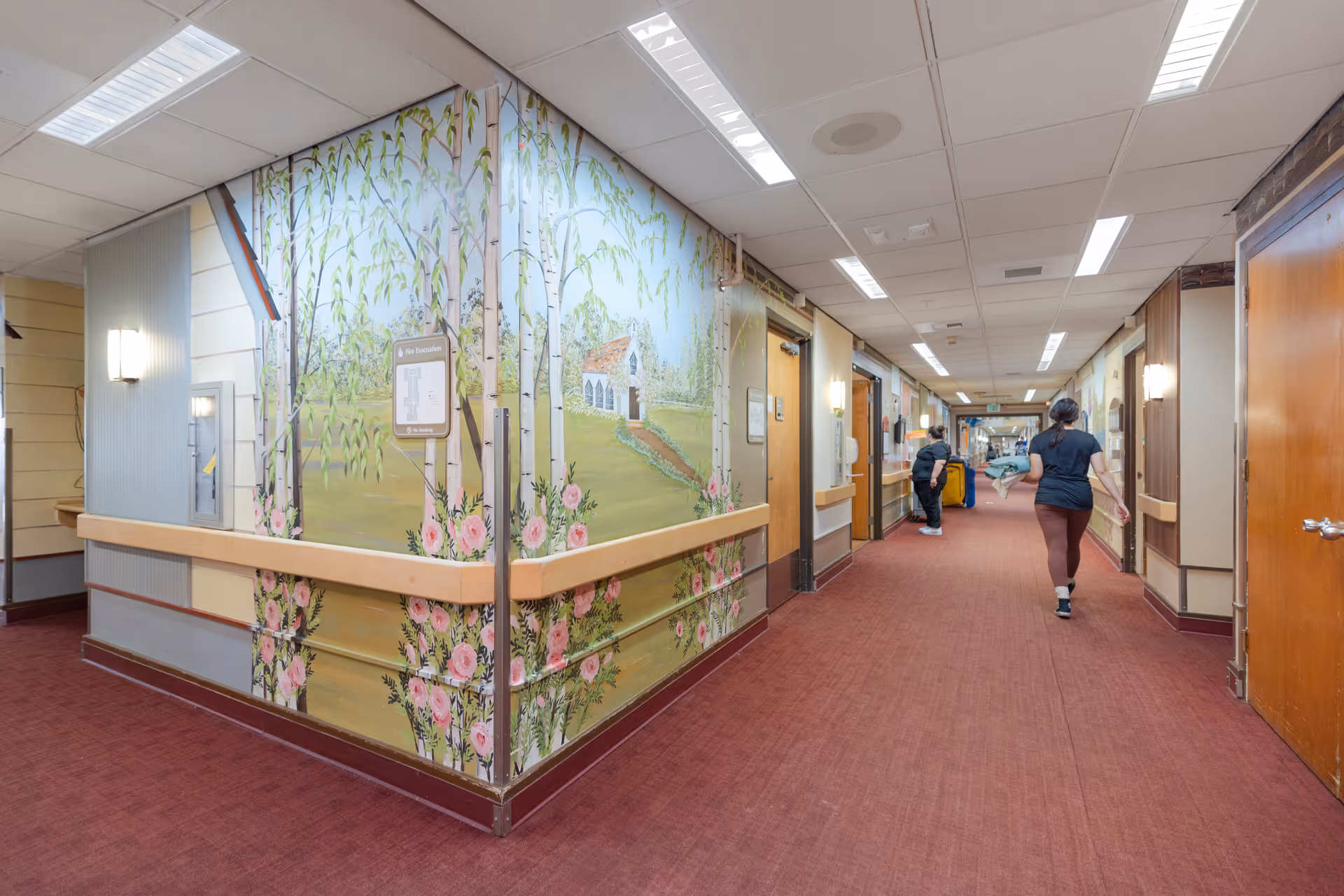 A long indoor hallway in a senior living facility with red carpet flooring and beige walls. The left corner wall features a mural of trees, flowers, and a small house. Two people are seen walking down the hallway, one carrying a stack of linens. The hallway is well-lit with ceiling lights and wall sconces.