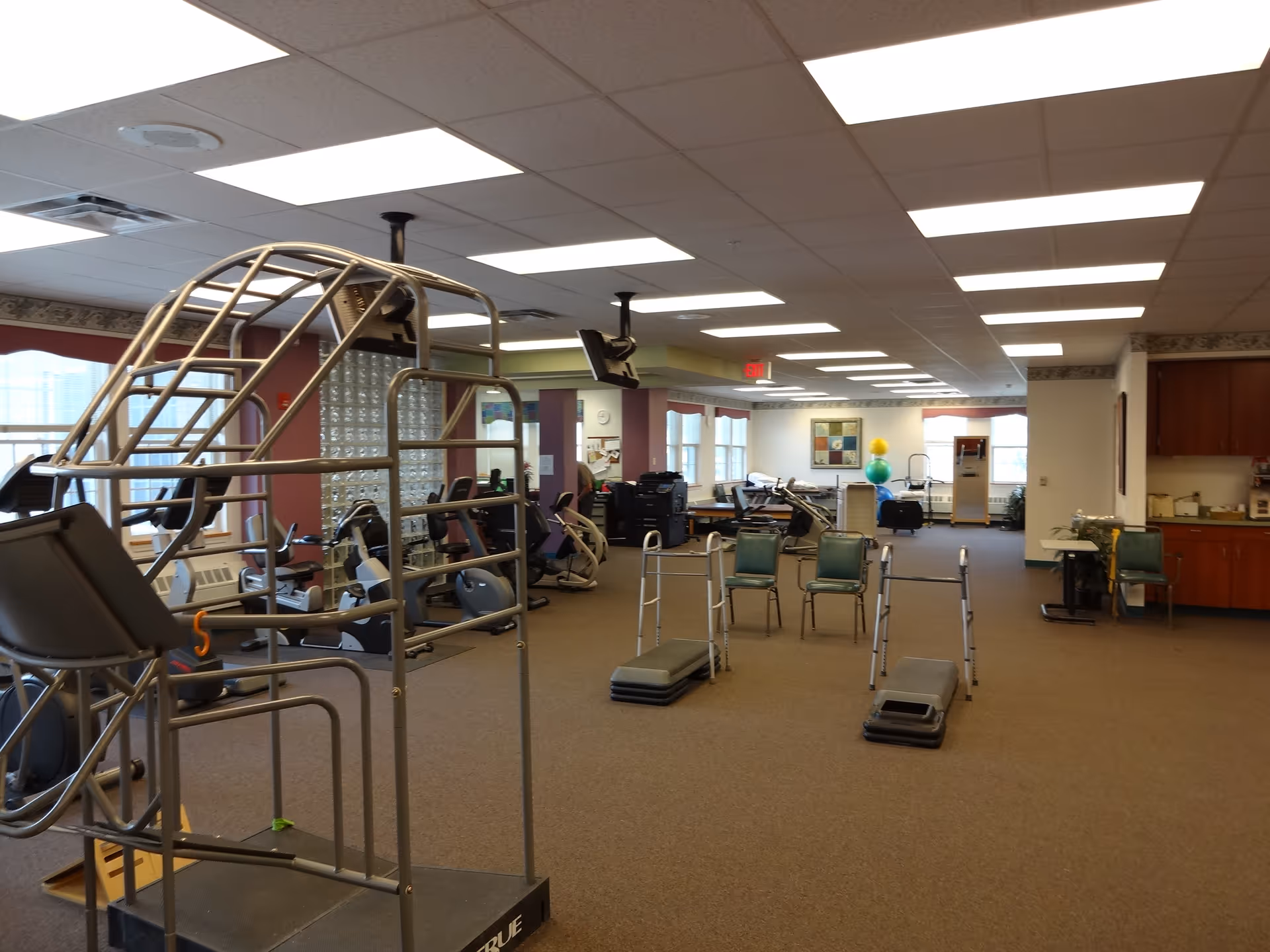 A spacious indoor exercise room with various fitness equipment including stationary bikes, walkers, step platforms, and a metal climbing frame. The room has carpeted floors, fluorescent ceiling lights, and large windows letting in natural light. There are chairs and a small kitchenette area with cabinets on the right side.