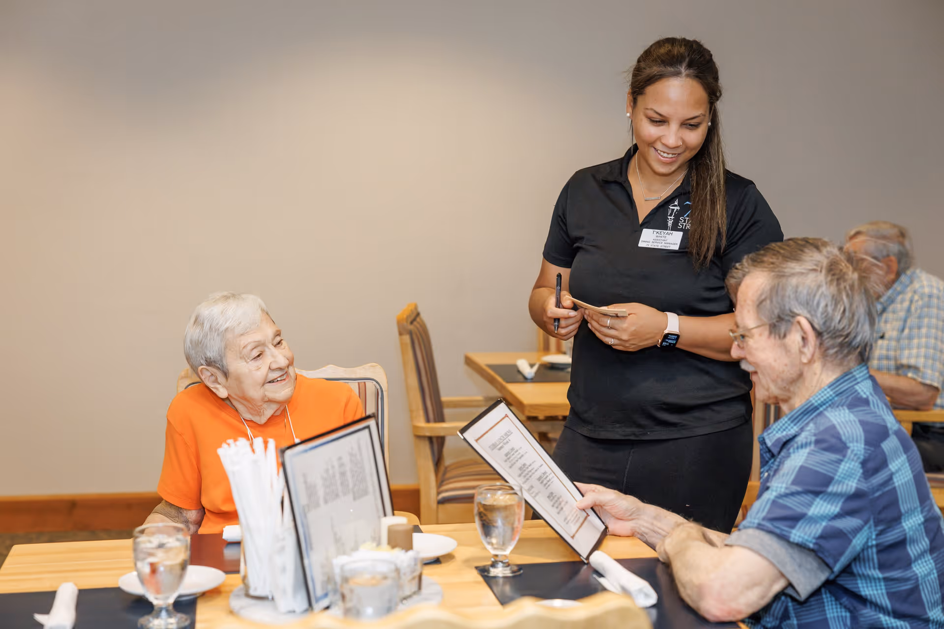 A waitress takes an order from two elderly customers seated at a dining table in a restaurant. The elderly woman in an orange shirt smiles at the waitress, while the elderly man in a blue plaid shirt looks at the menu. The table has glasses of water, menus, and utensils.