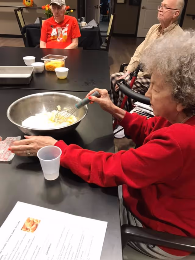 An elderly woman in a red sweater is whisking ingredients in a large metal bowl at a table. Two elderly men sit at the table in the background, one wearing a red AFC shirt and a cap, and the other in a beige shirt with a walker beside him. Various small bowls and a recipe sheet are also on the table.