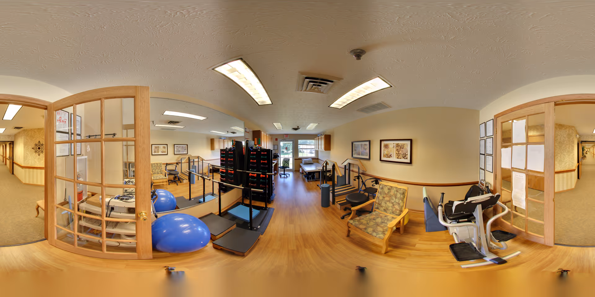Interior view of a senior living facility room equipped for physical therapy and exercise. The room features wooden flooring, exercise equipment including balance balls, parallel bars, and a stationary exercise bike. There are chairs with patterned cushions along the walls, framed artwork, and large windows letting in natural light. Double wooden doors with glass panels open into a hallway.