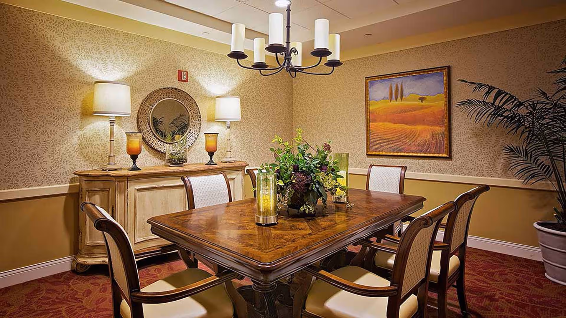 Formal dining room with a polished wooden table surrounded by chairs, a floral centerpiece, sideboard with lamps, and framed artwork.