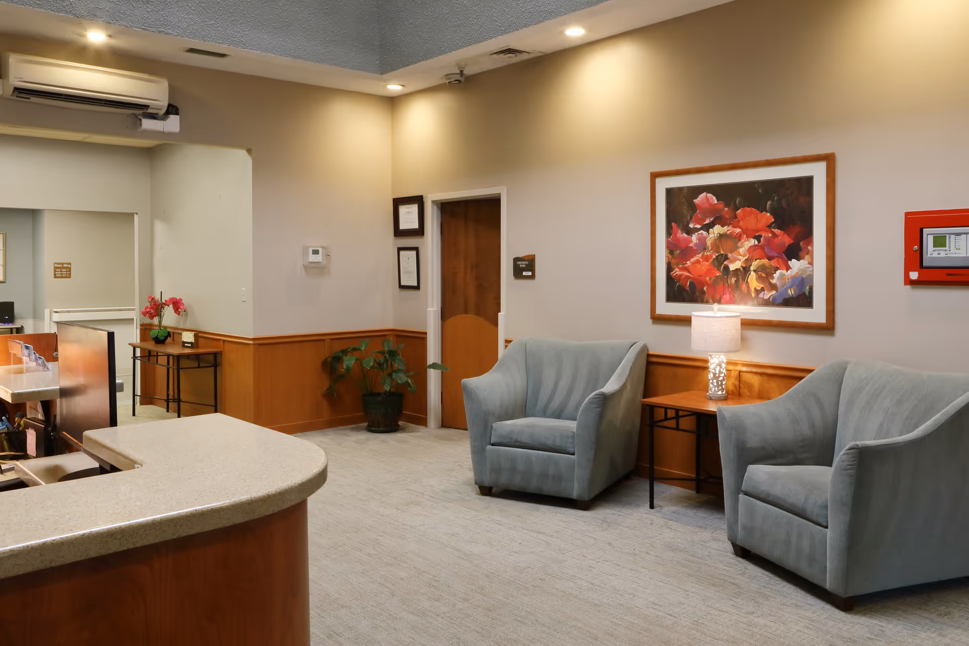 A cozy waiting area inside a facility with two light blue armchairs, a small wooden side table with a lamp, a colorful floral painting on the wall, a potted plant, and a reception desk with a curved countertop. The walls are beige with wood paneling on the lower half, and there is an air conditioning unit mounted near the ceiling.