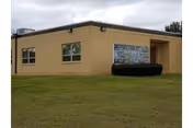 Single-story beige building with two windows and a small entrance area, surrounded by grass under a cloudy sky. A sign on the building reads Gilmer Nursing & Rehabilitation.