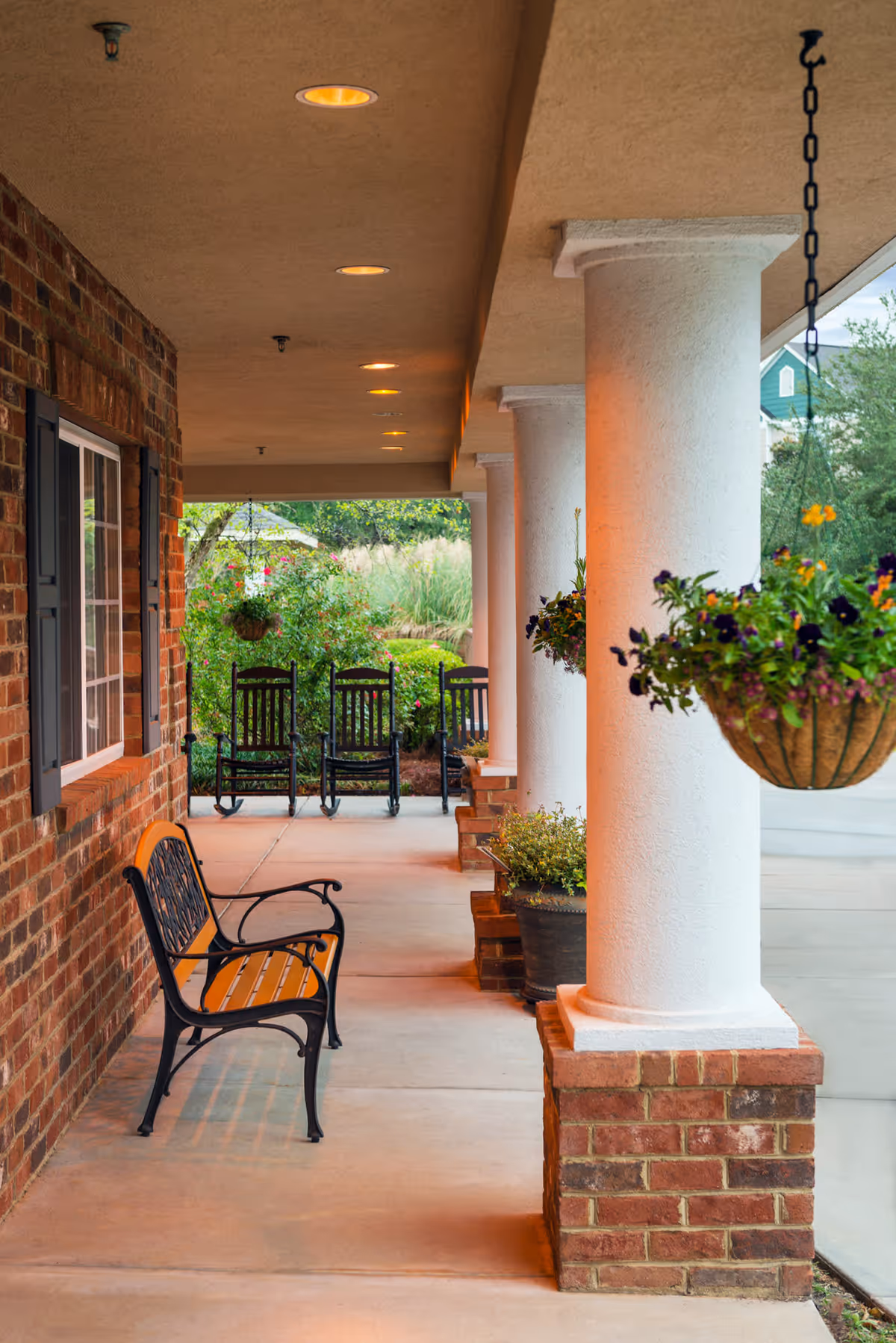 Covered outdoor porch area with white columns and brick bases, featuring a wooden bench with black metal frame in the foreground and three black rocking chairs in the background. Hanging flower baskets and potted plants decorate the space, with greenery and trees visible beyond the porch.