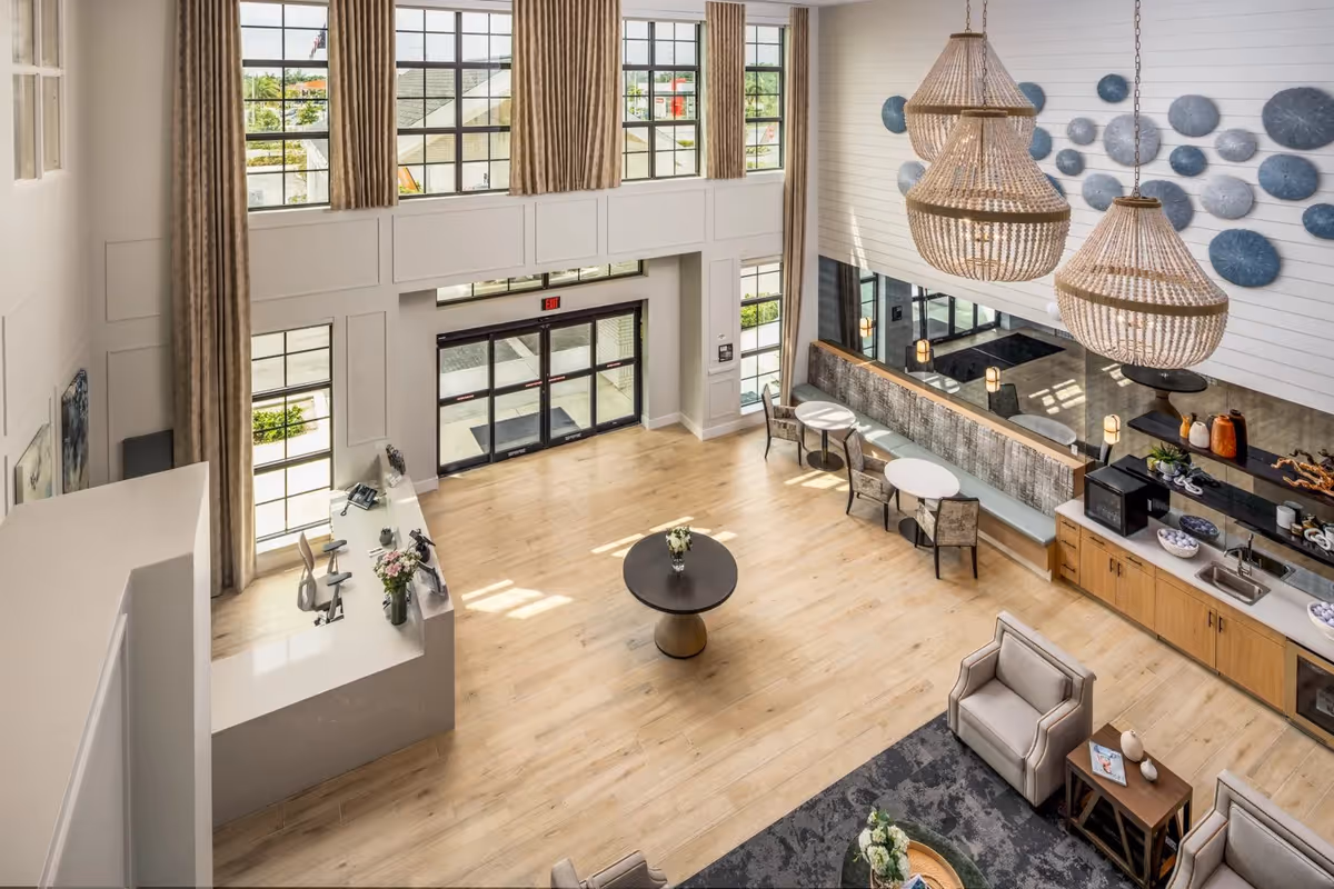 Spacious and bright senior living facility lobby with large windows, a reception desk, seating area with armchairs and tables, decorative wall art, and two large hanging chandeliers.