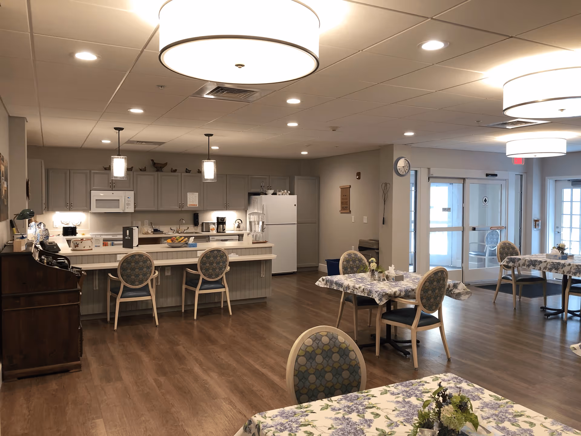 Open communal dining area with several tables and chairs in front of a small kitchen and serving counter under pendant lights.