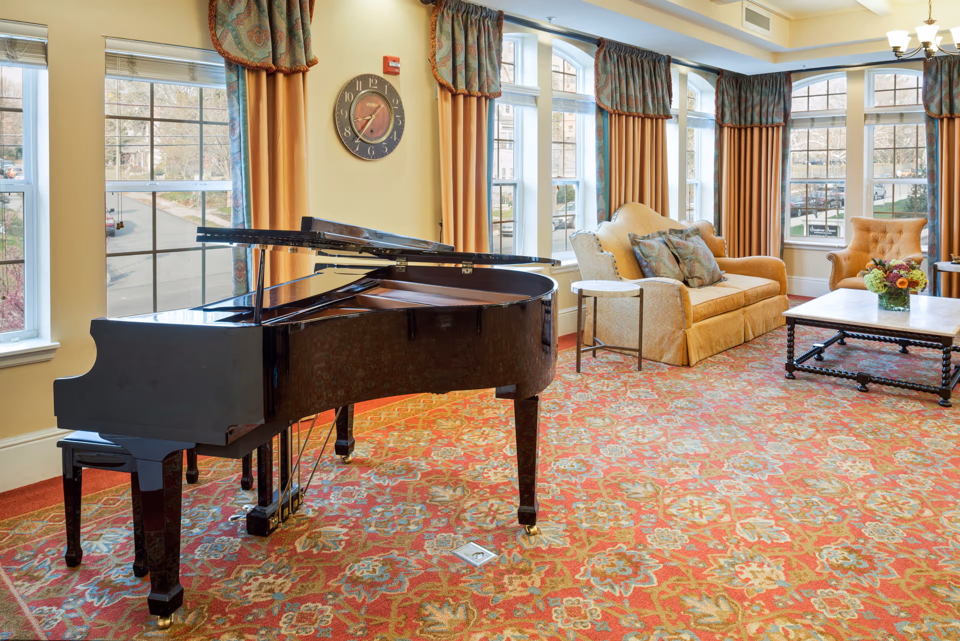 A bright and elegant living room with large windows covered by patterned curtains, a black grand piano with an open lid, a beige sofa with decorative pillows, an armchair, a coffee table with a flower arrangement, and a colorful patterned carpet.