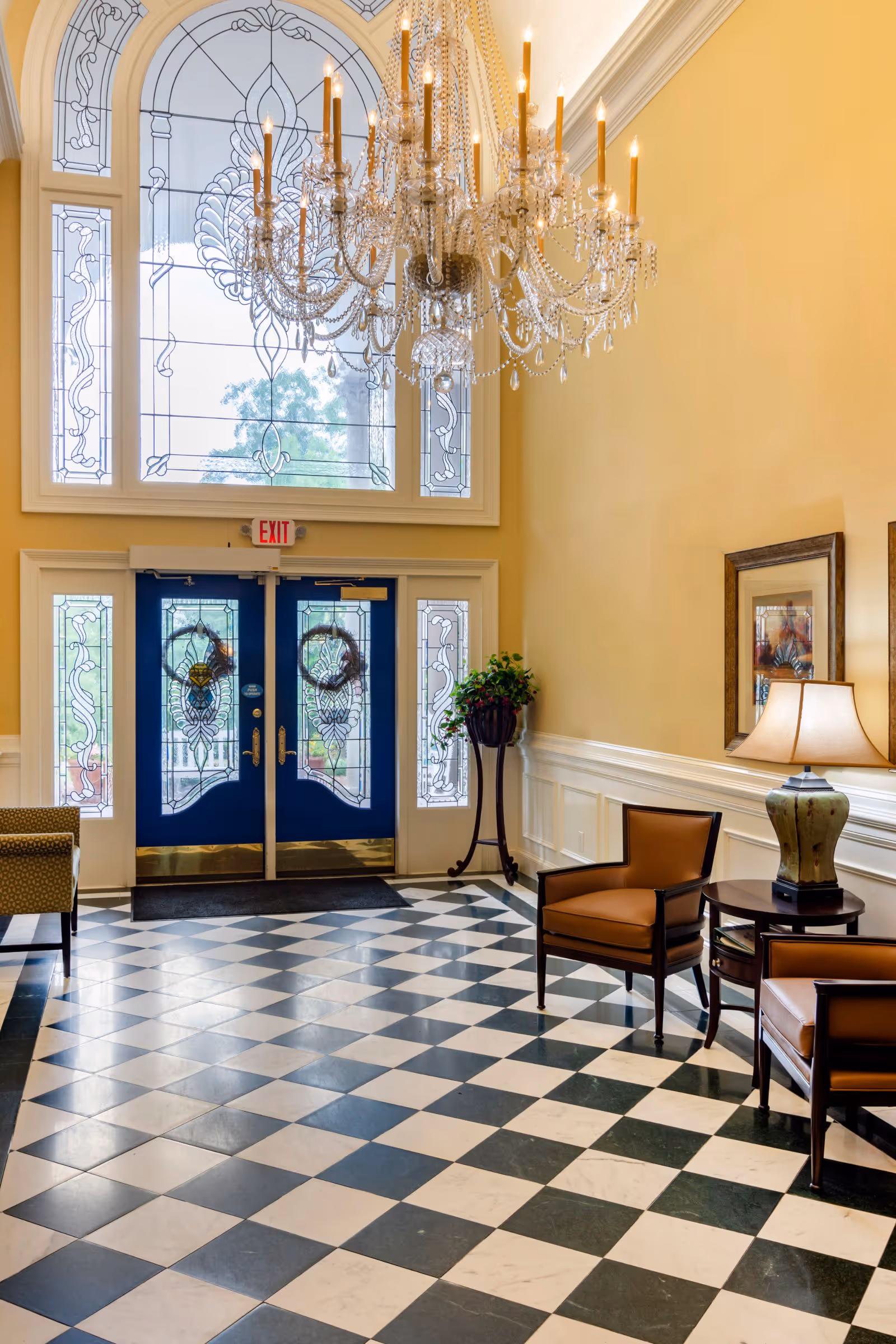 A bright and elegant lobby area with a large crystal chandelier hanging from a high ceiling. The floor features a black and white checkered pattern. There are two blue double doors with decorative glass panels and an exit sign above them. To the right, there are two brown leather chairs with a small table and a lamp between them. A potted plant stands near the door, and framed artwork hangs on the wall.