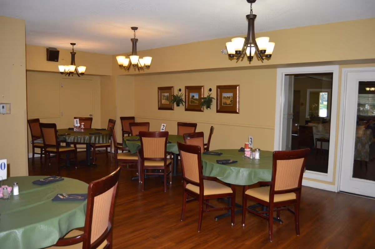 Interior view of a dining room with several round tables covered with green tablecloths and set with napkins and utensils. Each table has a number card and condiments. The room has wooden chairs with cushioned seats, yellow walls adorned with framed pictures and plants, and three ceiling light fixtures. There is a door and a large window on one side of the room.