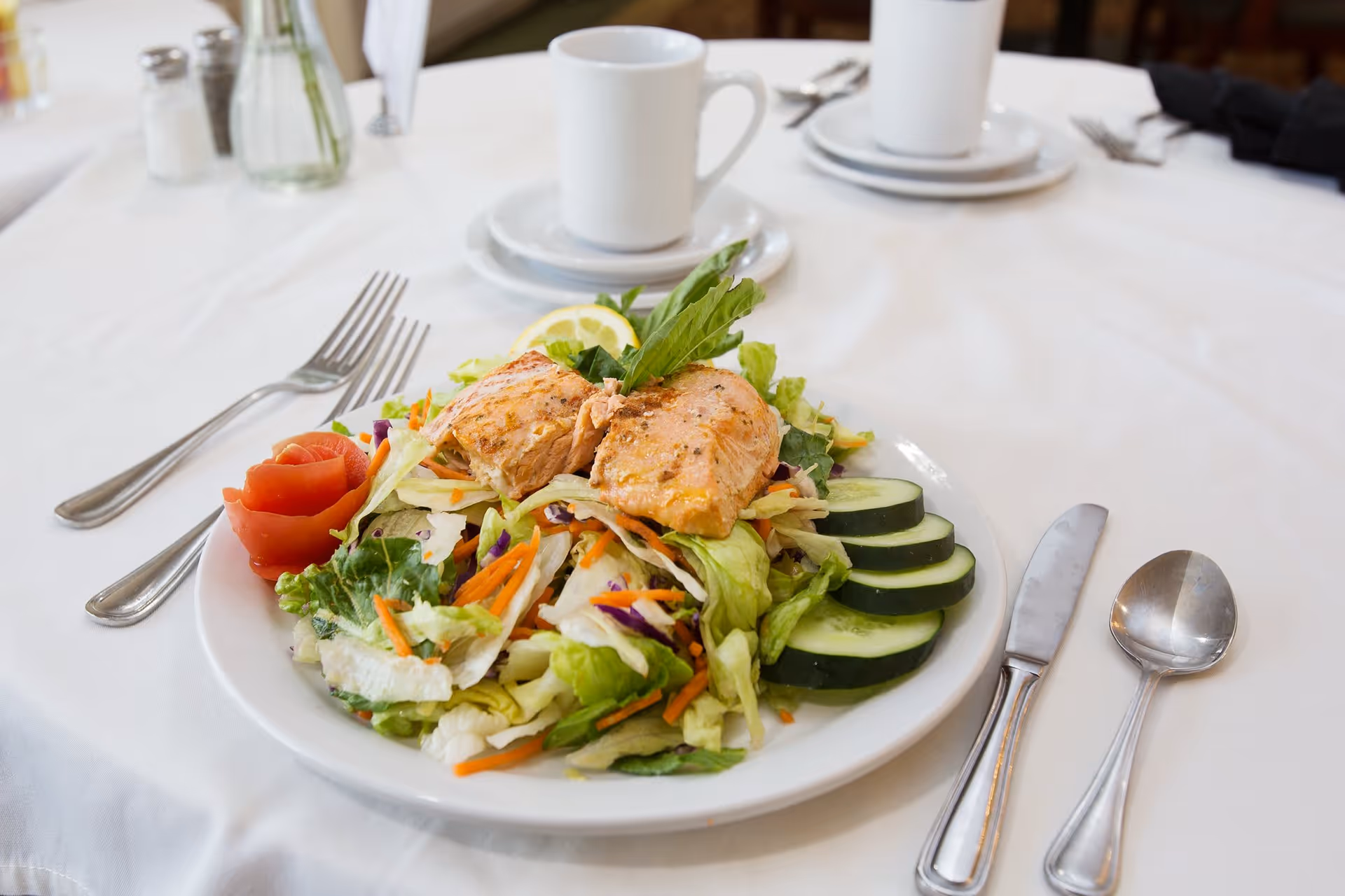 A plate of fresh salad with grilled salmon, sliced cucumbers, and a tomato rose garnish on a white tablecloth. There are utensils, a white coffee cup on a saucer, and salt and pepper shakers in the background.