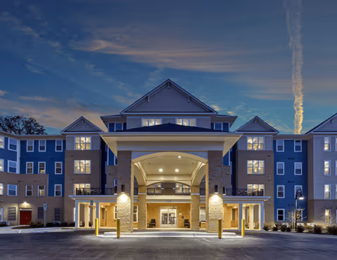 Front exterior view of a large, multi-story senior living facility named Pointe View at Aspen Hill during dusk, with well-lit entrance and symmetrical architecture featuring multiple windows and a covered drop-off area.