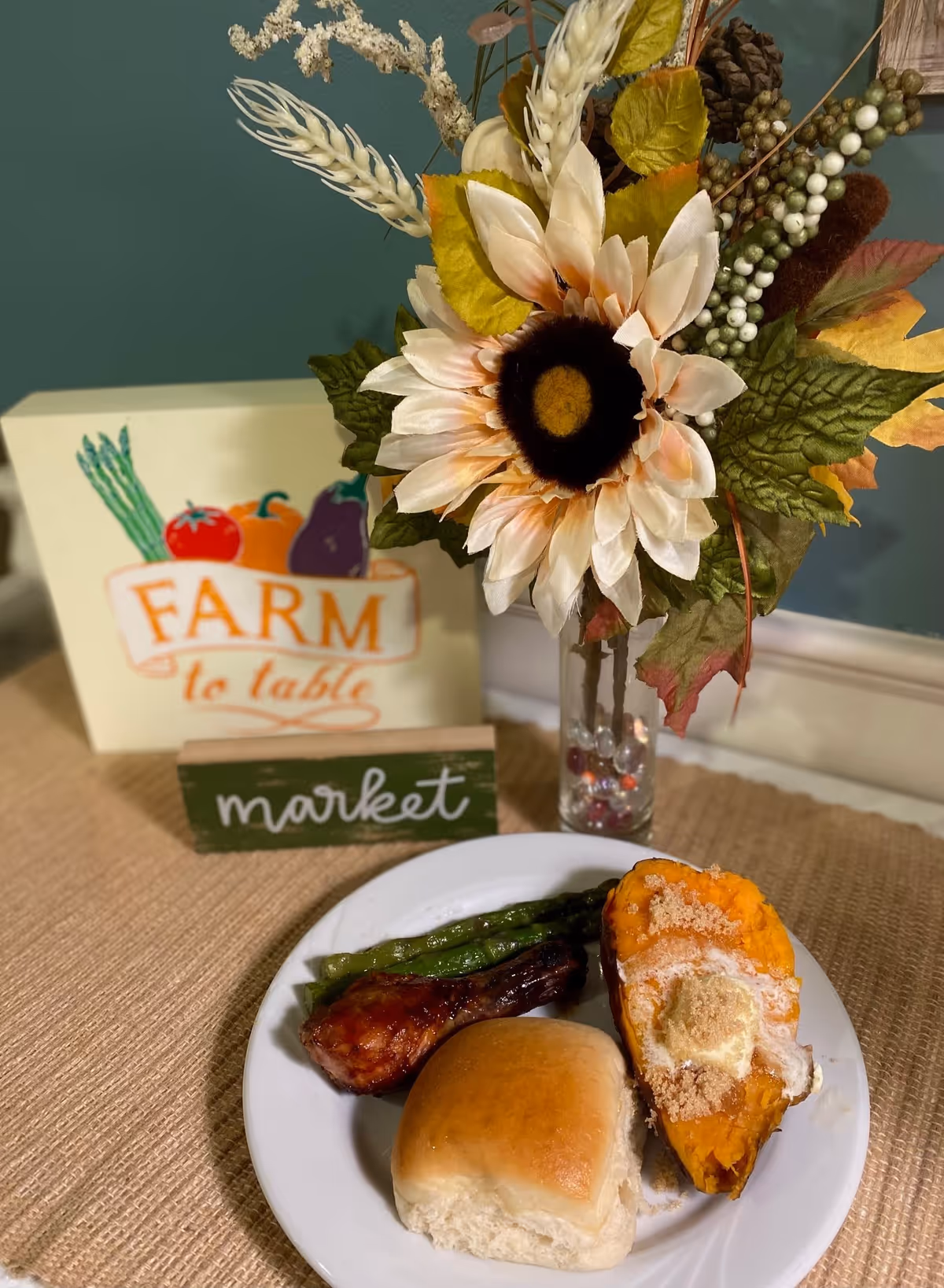 A plate with a baked sweet potato topped with butter and brown sugar, a dinner roll, a grilled chicken drumstick, and asparagus. Behind the plate is a vase with a large artificial sunflower and other decorative foliage. There are two small signs in the background, one reading 'FARM to table' with illustrations of vegetables, and the other reading 'market'.