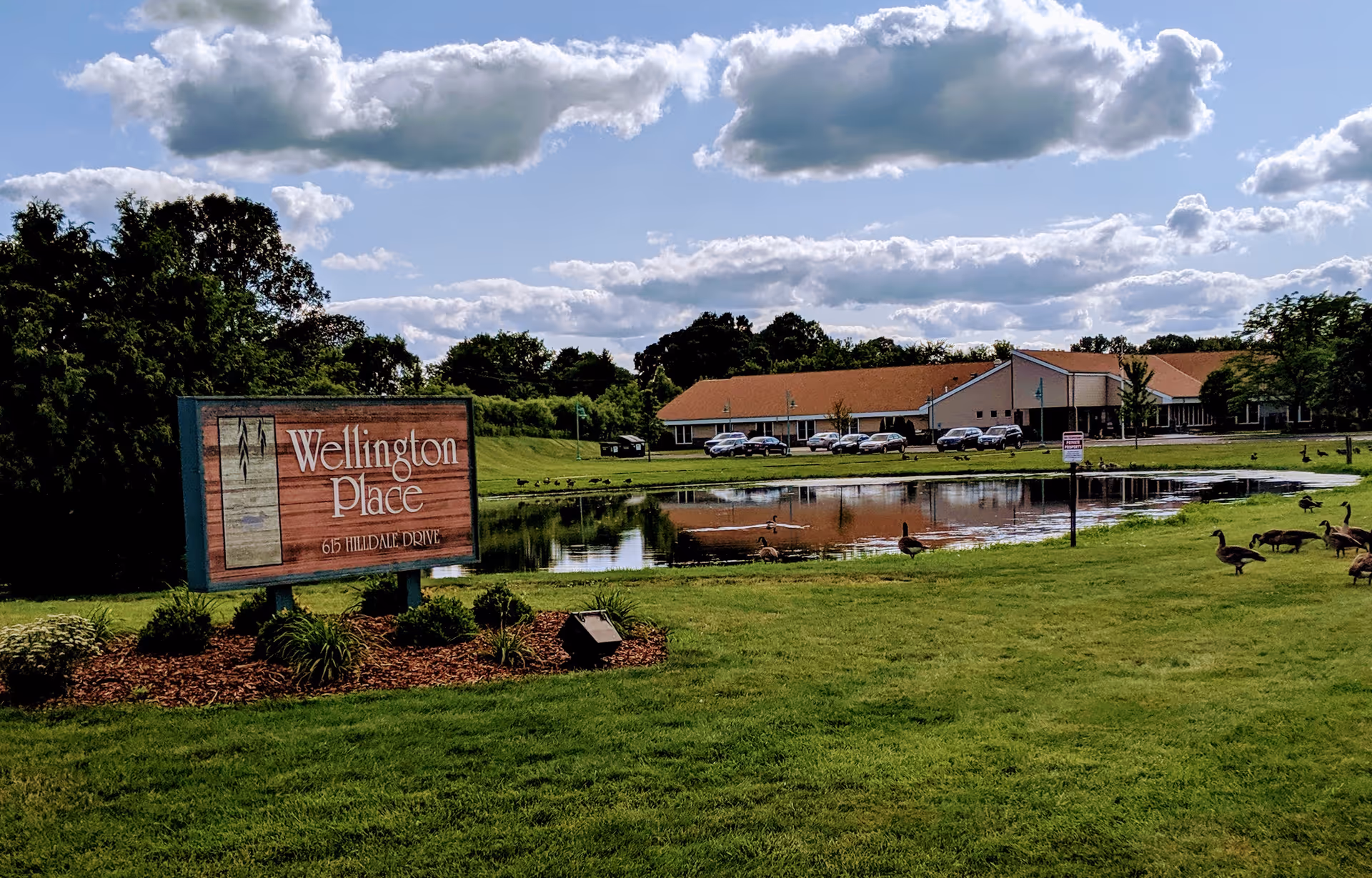 A Wellington Place entrance sign on a landscaped lawn by a pond with the facility building and parked cars in the background under a partly cloudy sky.