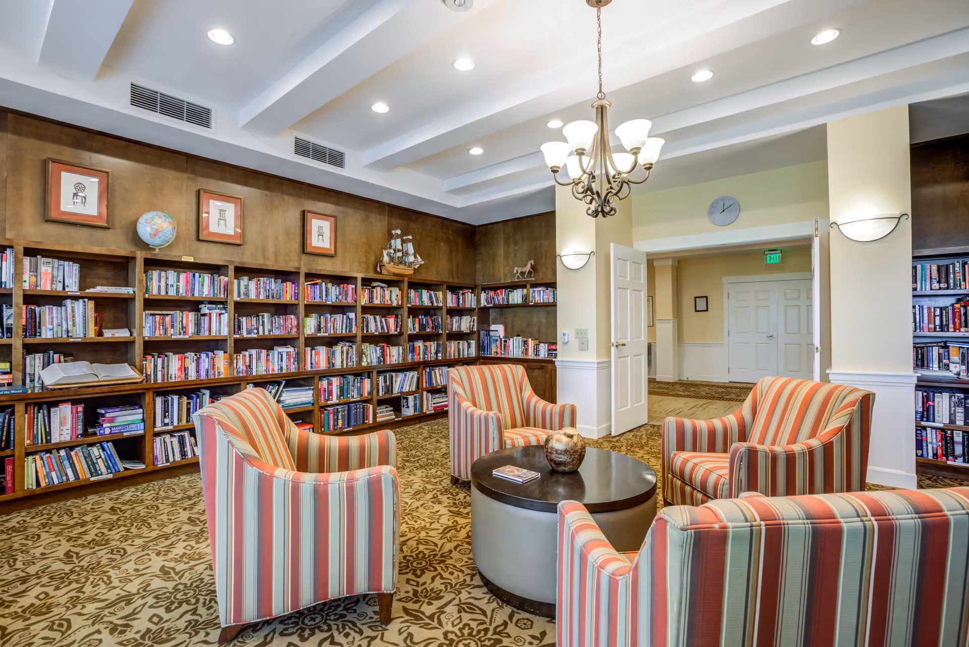 A cozy library room with wooden bookshelves filled with books along the walls. Four striped armchairs surround a round coffee table in the center. The room has a patterned carpet, a chandelier hanging from the ceiling, and framed pictures and decorative items on the shelves.