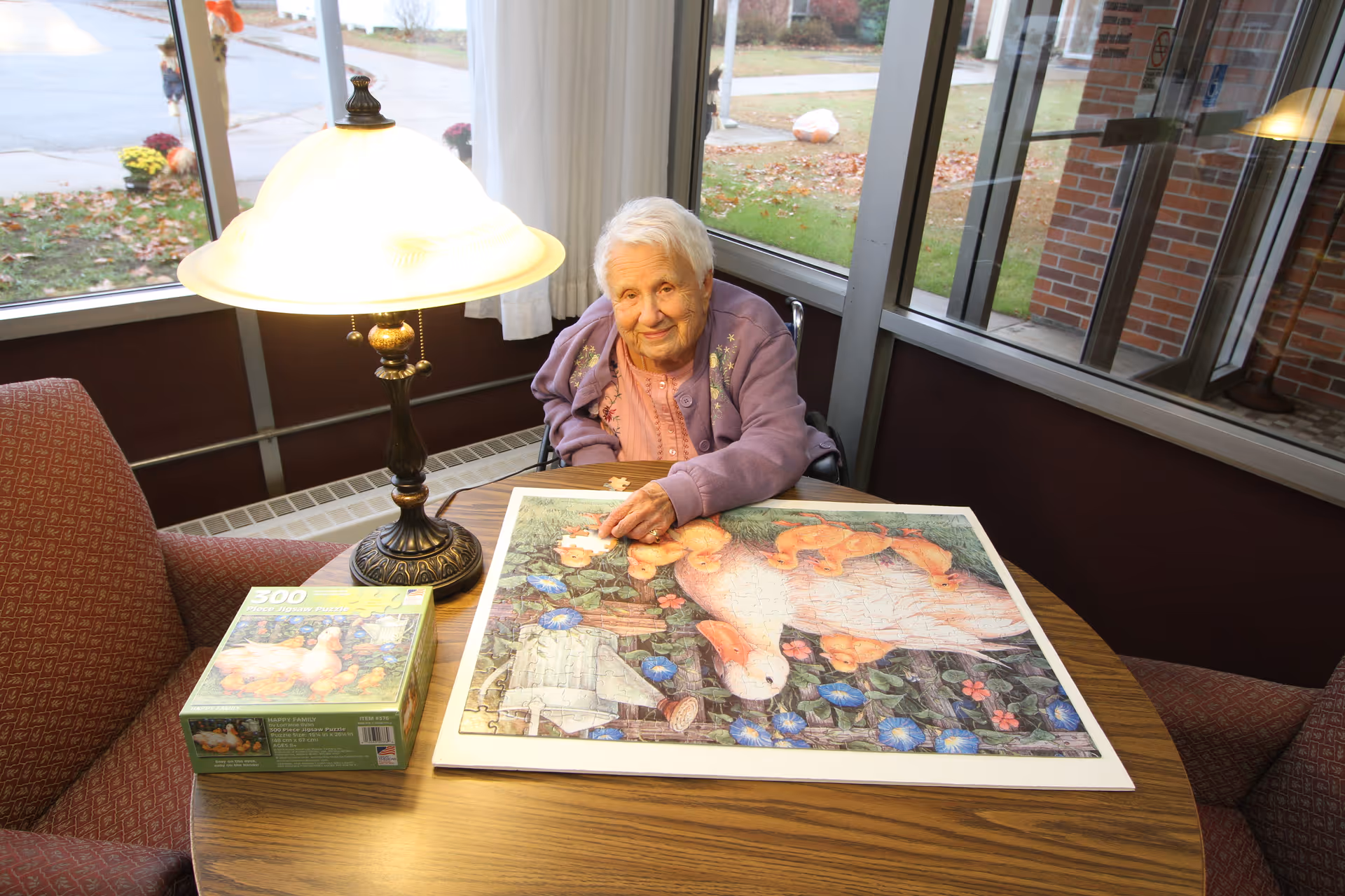 An elderly woman sitting at a round wooden table in a cozy room with large windows, working on a completed jigsaw puzzle featuring ducks and flowers. A table lamp with a white shade is lit beside her, and the puzzle box is also on the table.